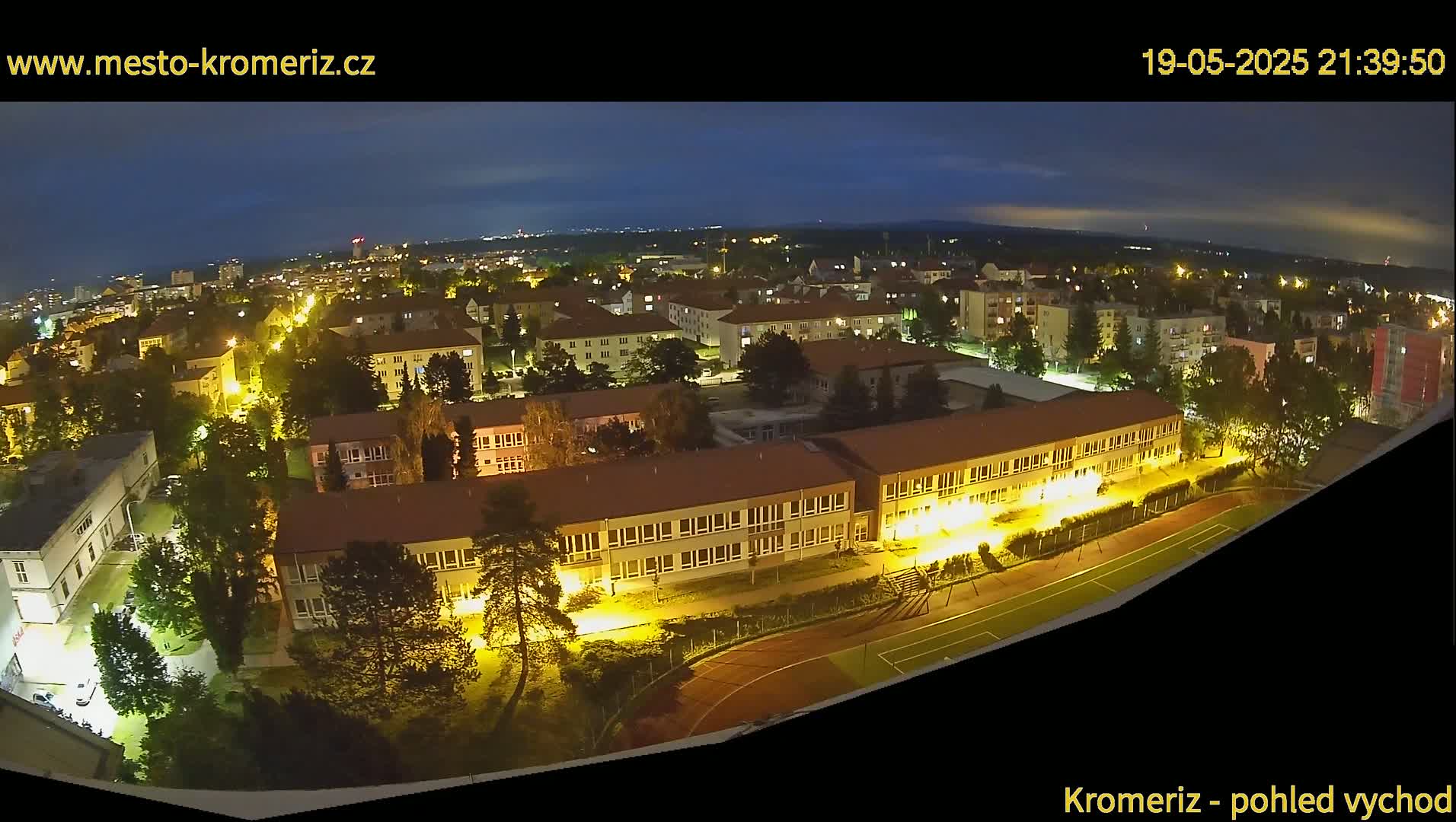 A nighttime aerial view shows a town illuminated with streetlights, featuring a large school building and athletic field.
