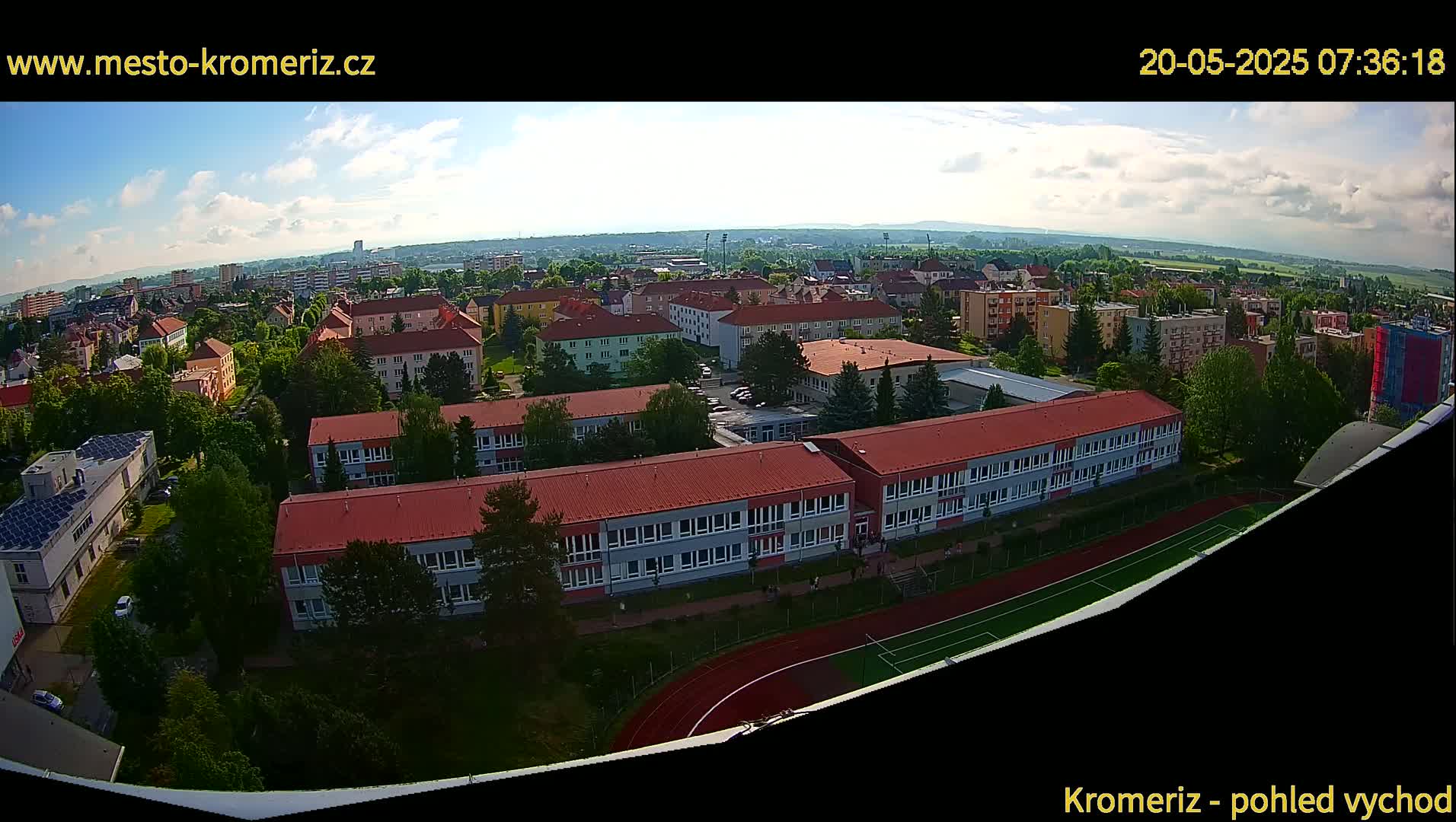 An aerial view of a town on a partly cloudy day shows several buildings, including a large school with a red roof and a running track, surrounded by trees and other structures.