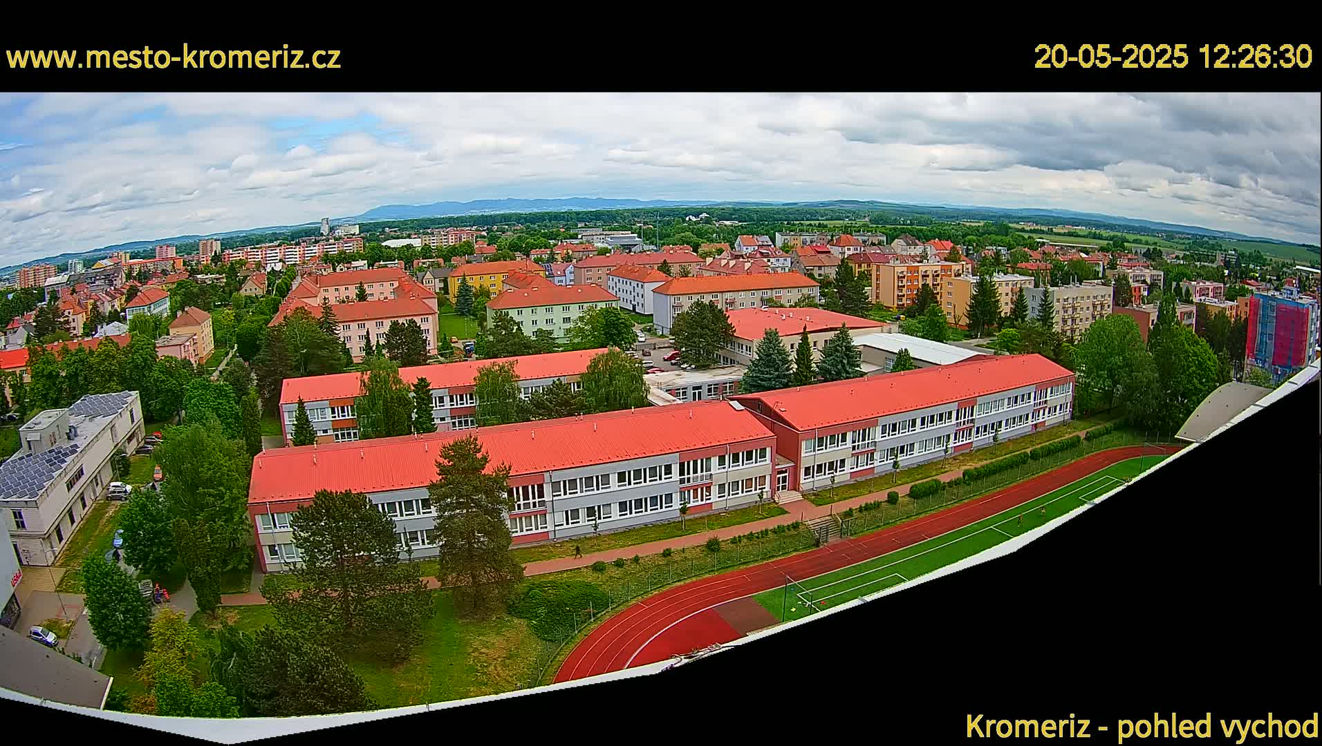 An aerial view shows a school complex with red roofs and a running track, situated in a town under a partly cloudy sky.