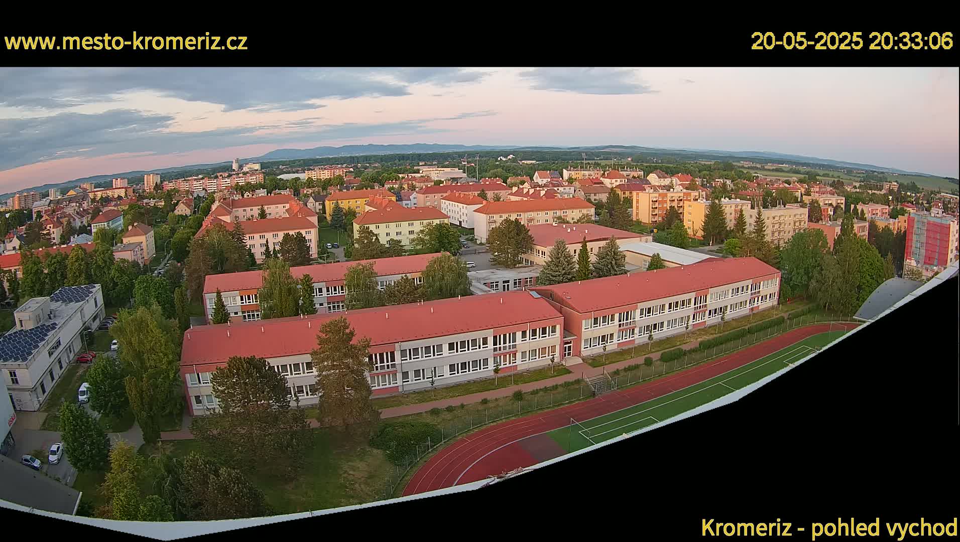 An aerial view shows a residential area with several buildings, including a large school with a running track and a soccer field, during the evening under a mostly clear sky.
