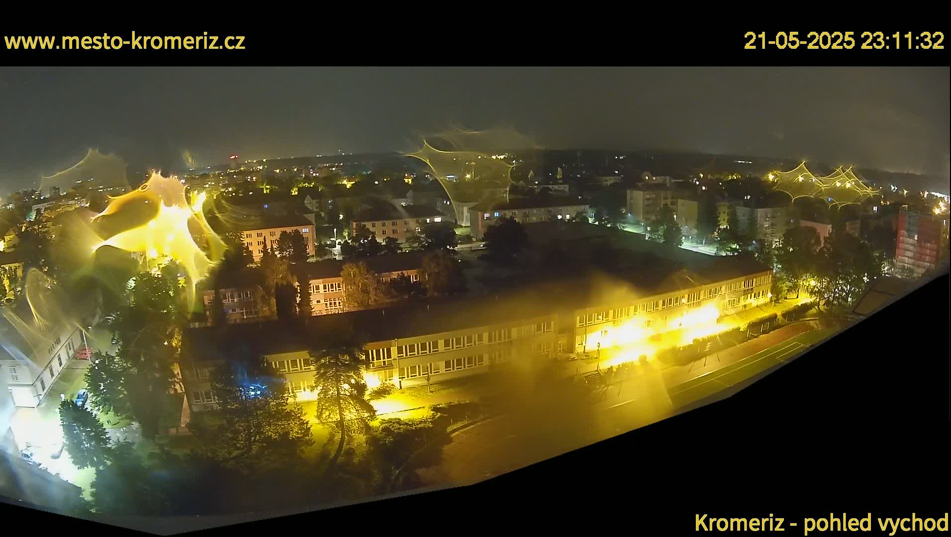 A nighttime aerial view of a town shows a large building illuminated with bright yellow light, surrounded by smaller buildings and trees under a dark, clear sky.