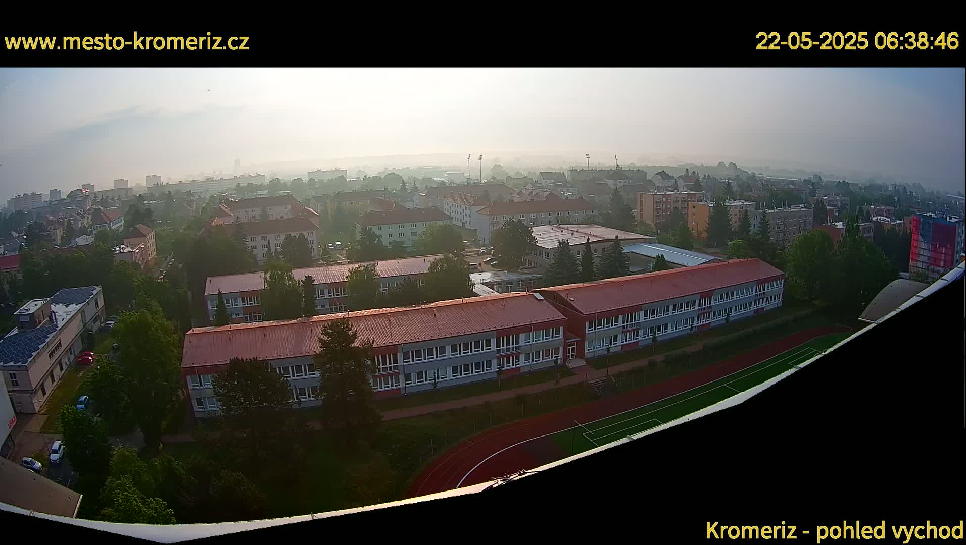 A hazy sunrise reveals a sprawling cityscape of low-rise buildings, a school, and athletic fields.