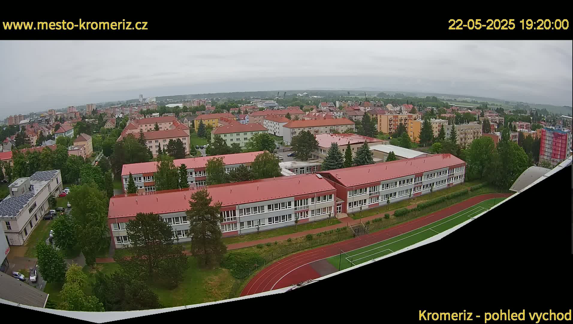 An aerial view shows a school complex with red roofs, surrounded by trees and a running track, under an overcast sky.