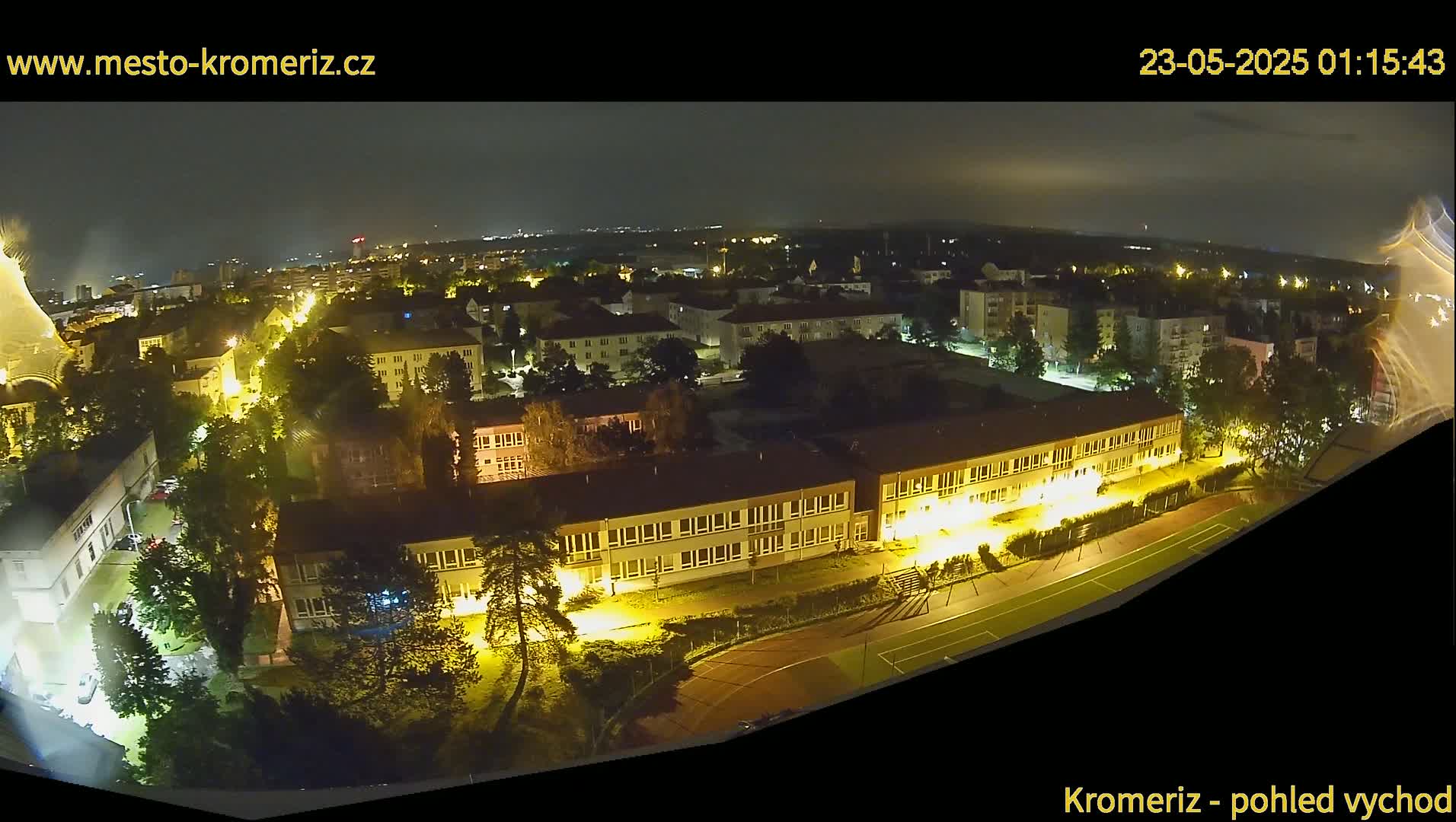 A nighttime aerial view of a town shows buildings with illuminated windows, trees, and a running track.