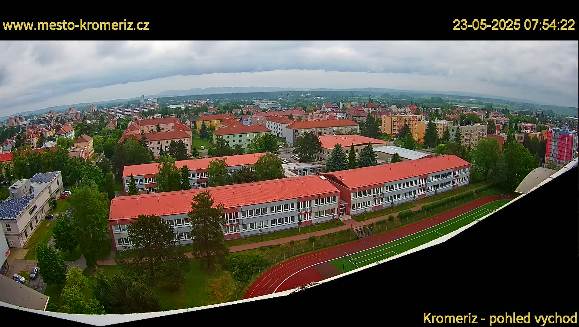 An aerial view shows a town under a cloudy sky, with several buildings, including a large school with a red roof and a running track, visible.