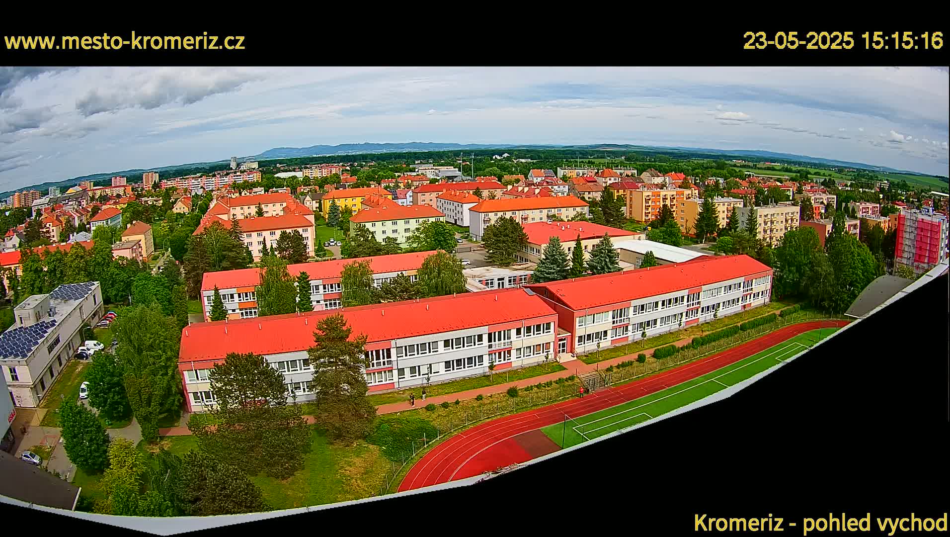 An aerial view shows a town under partly cloudy skies, with a large school building and a running track prominently featured in the foreground.