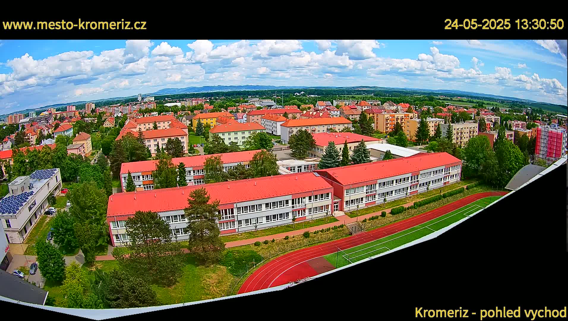 An aerial view shows a large school complex with red roofs surrounded by residential buildings under a partly cloudy sky.