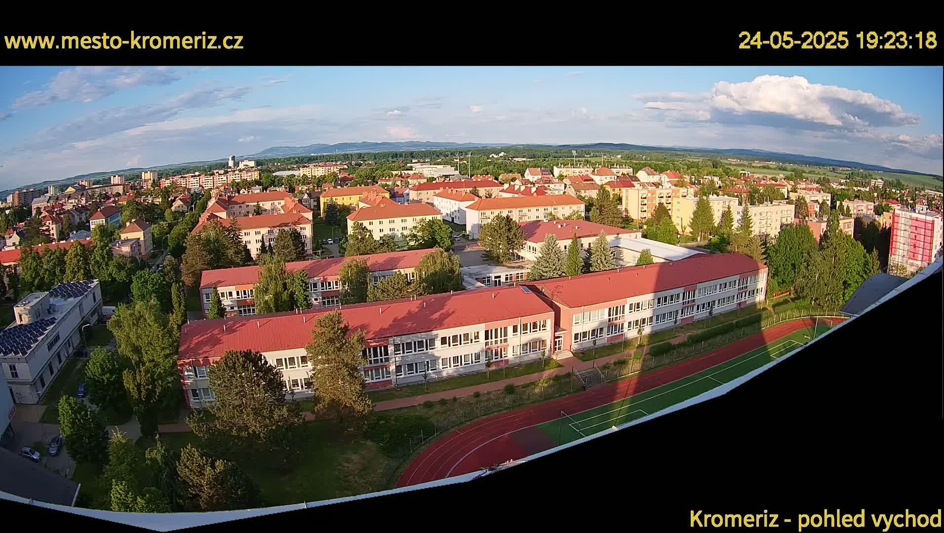 An aerial view of a town under partly cloudy skies at sunset, showing a large school building, surrounding residential buildings, and a running track.