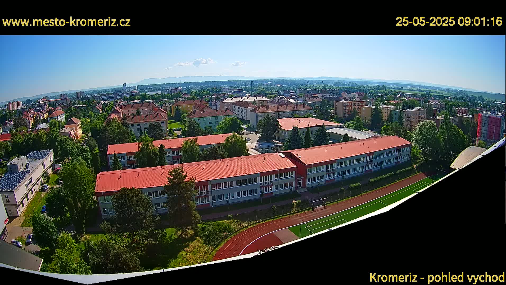 An aerial view of a town on a clear, sunny day shows several buildings, including a large school with a red roof and a running track, surrounded by trees and other structures.
