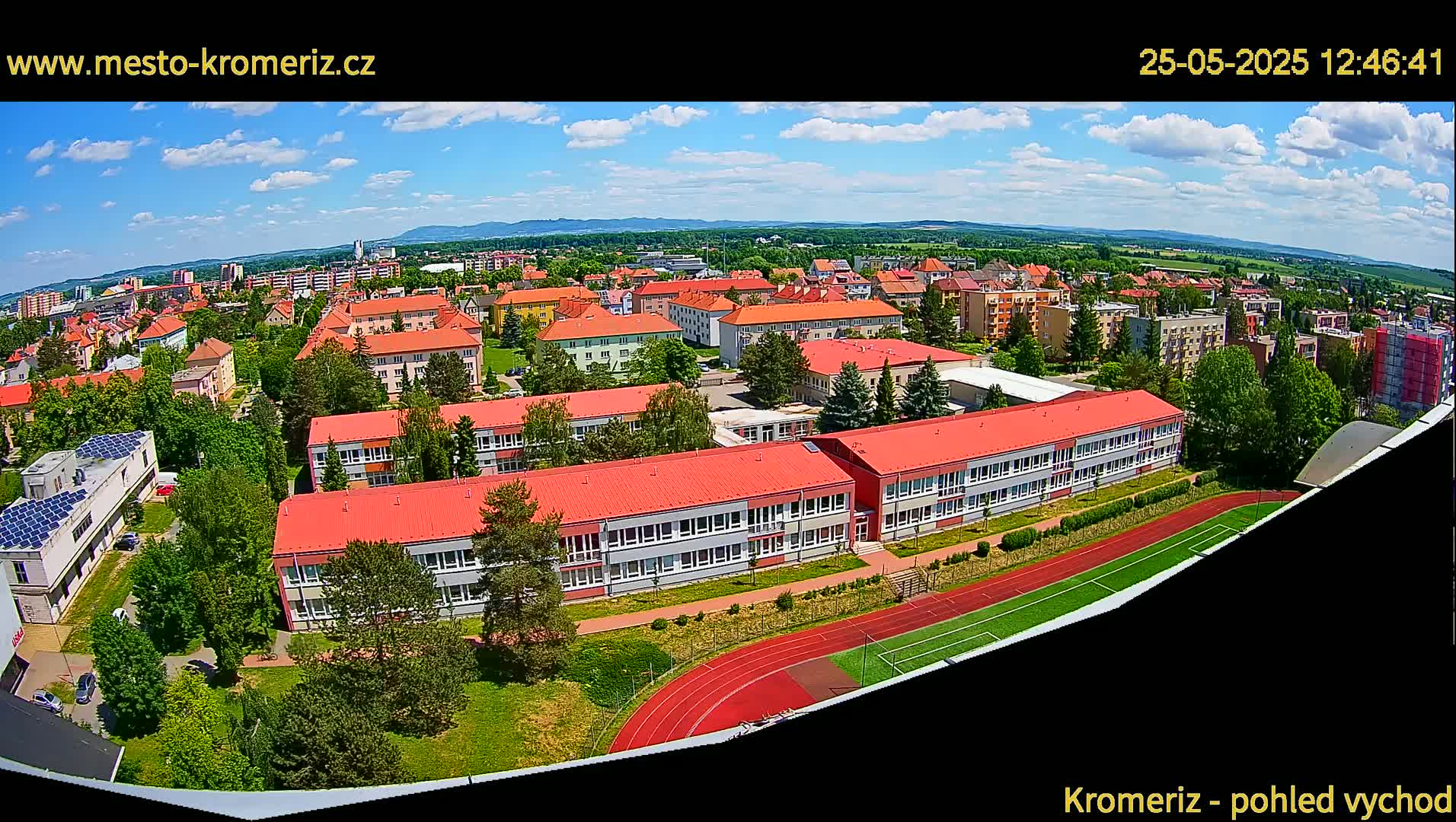 A high-angle, wide shot on a partly sunny day shows a large school building with a red roof and surrounding green space, including a running track, situated in a residential area with low-rise buildings extending to a distant horizon under a partly cloudy sky.