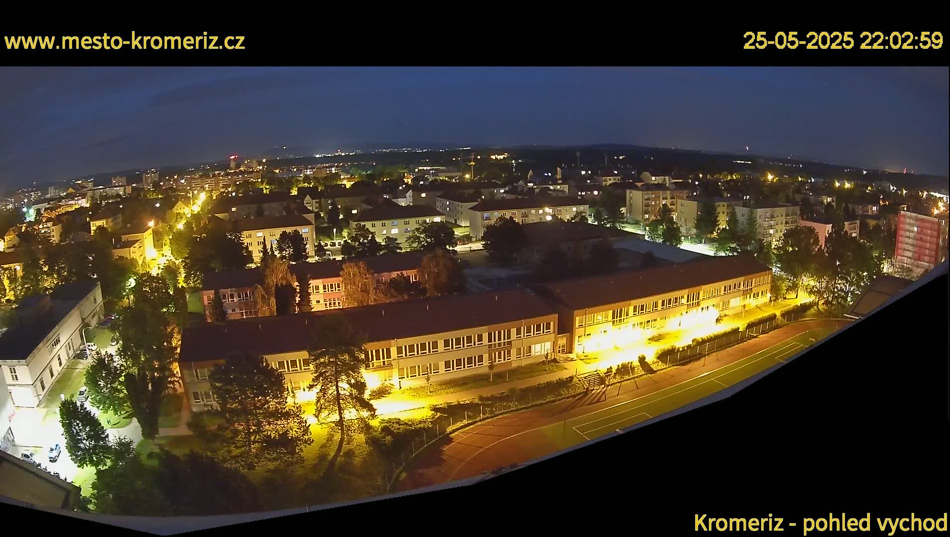 A nighttime aerial view shows a town's illuminated buildings and streets, with a school and athletic field prominent in the foreground, under a dark sky.