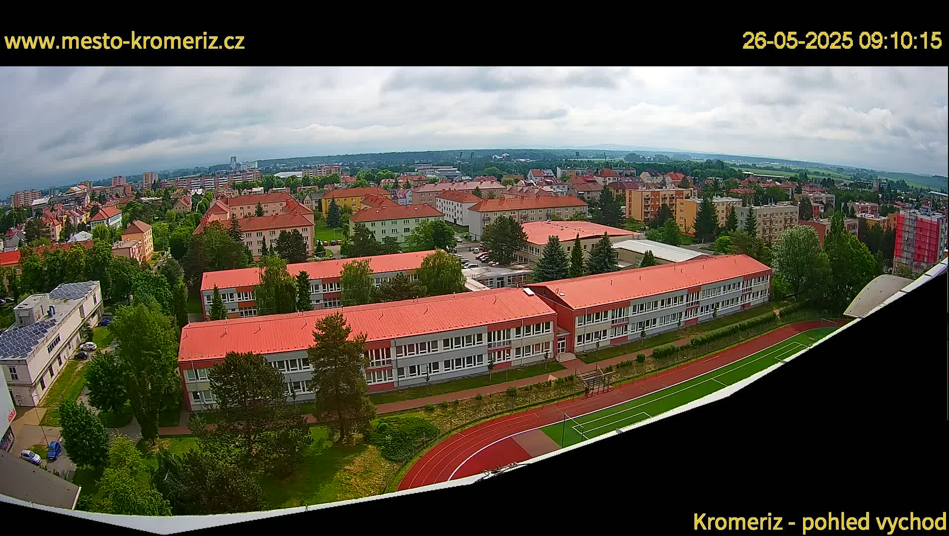 An aerial view shows a school complex with red roofs, surrounded by green trees and a running track, under a partly cloudy sky.