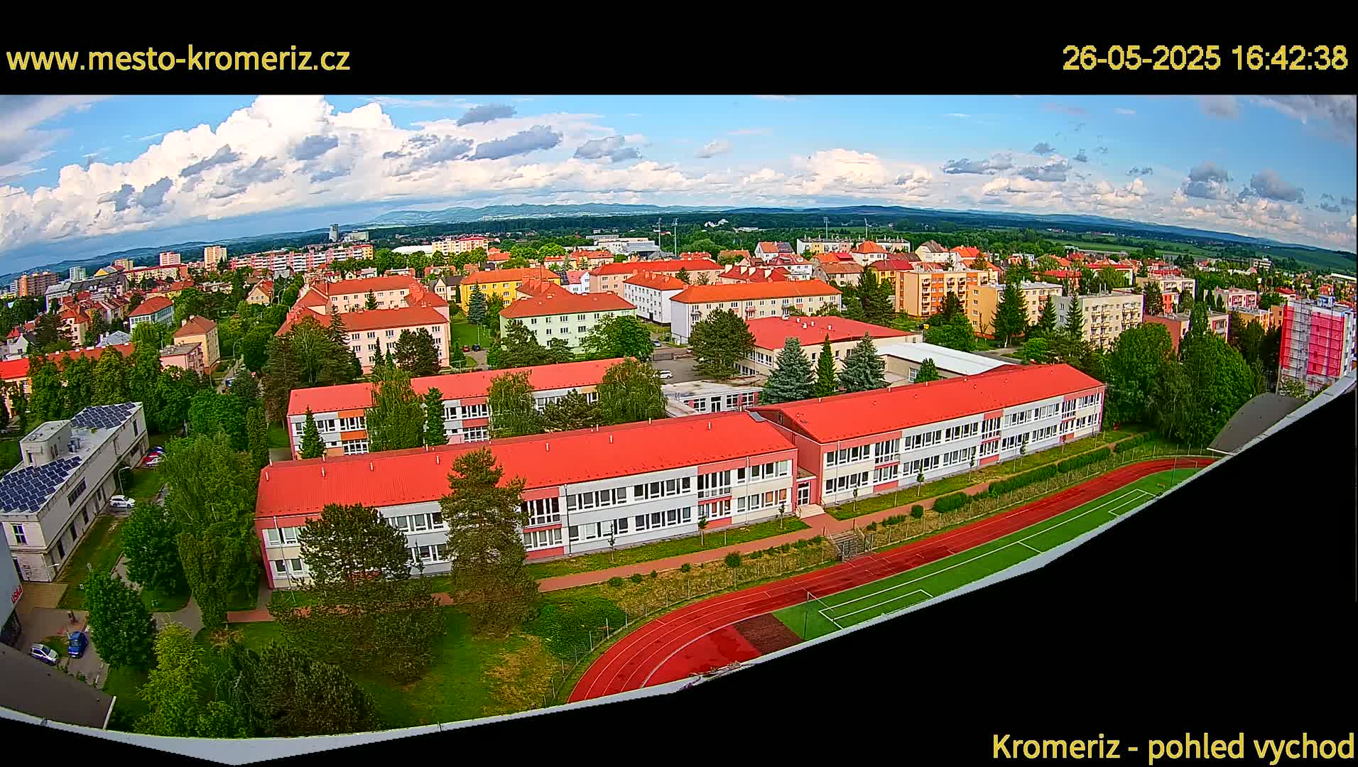 An aerial view shows a partly cloudy day over a town with numerous red-roofed buildings, a school, a running track, and a soccer field.