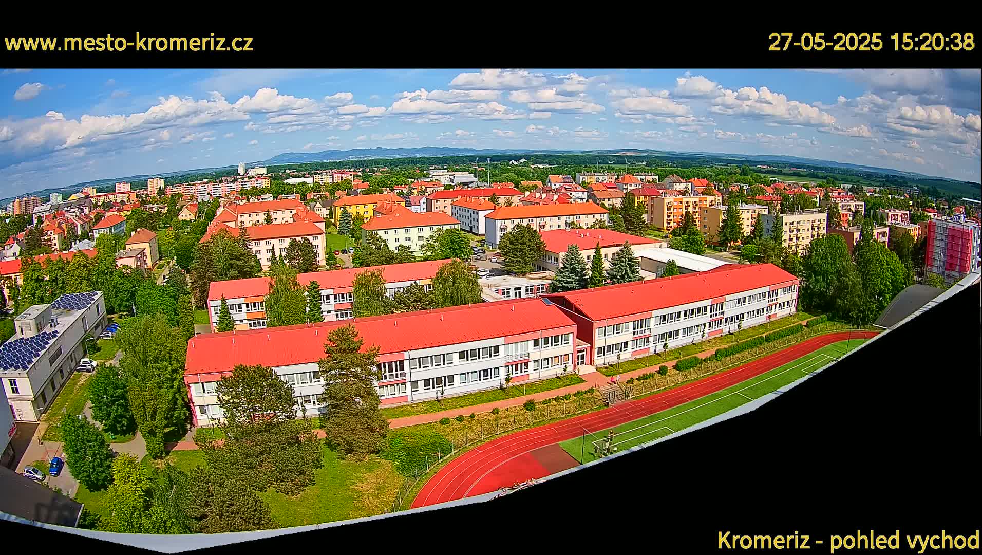 A partly cloudy day reveals a sprawling town with many red-roofed buildings, a large school, and a running track.