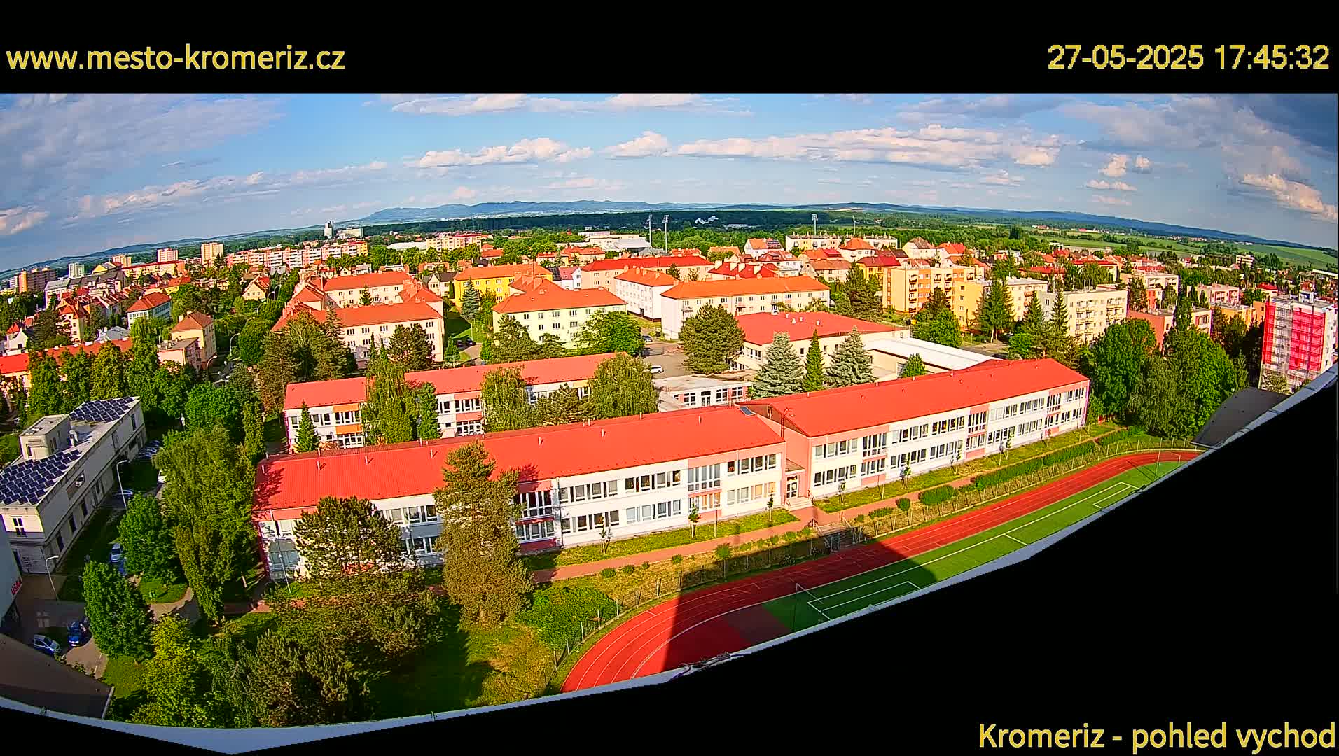 A partly sunny day reveals an aerial view of a town, including a large school building with red roofs, a running track, and surrounding residential buildings.