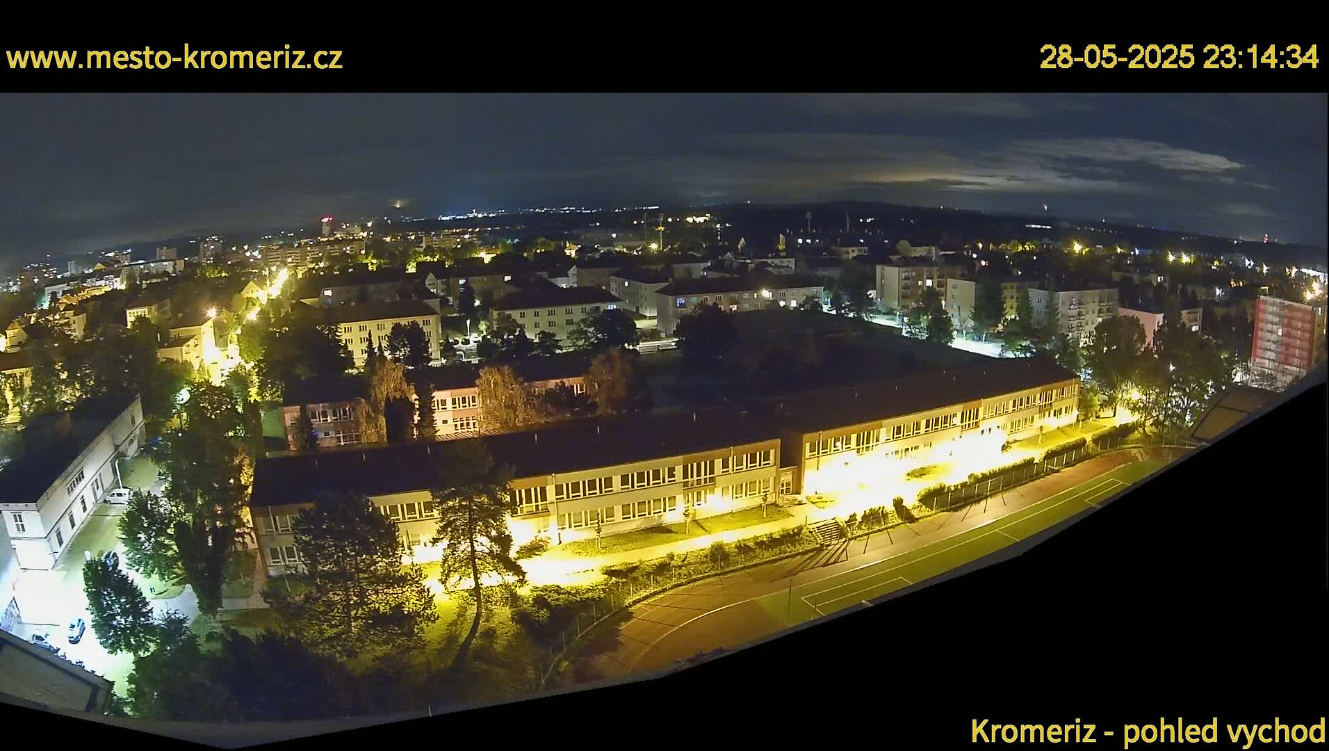 A nighttime aerial view of a town with illuminated buildings and a running track, under a partly cloudy sky.