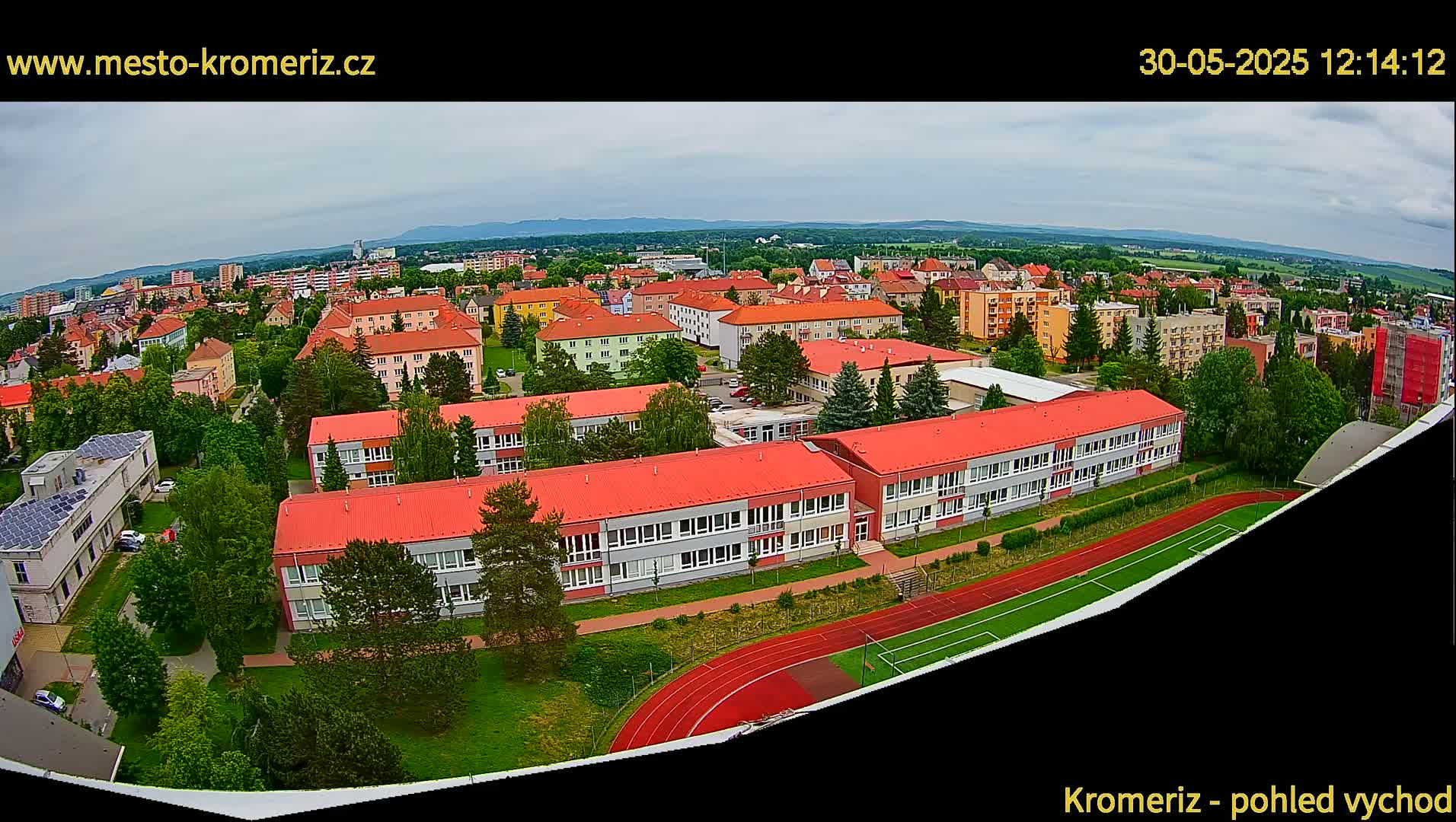 An aerial view shows a school complex with red roofs, surrounded by other buildings and greenery, under a partly cloudy sky.