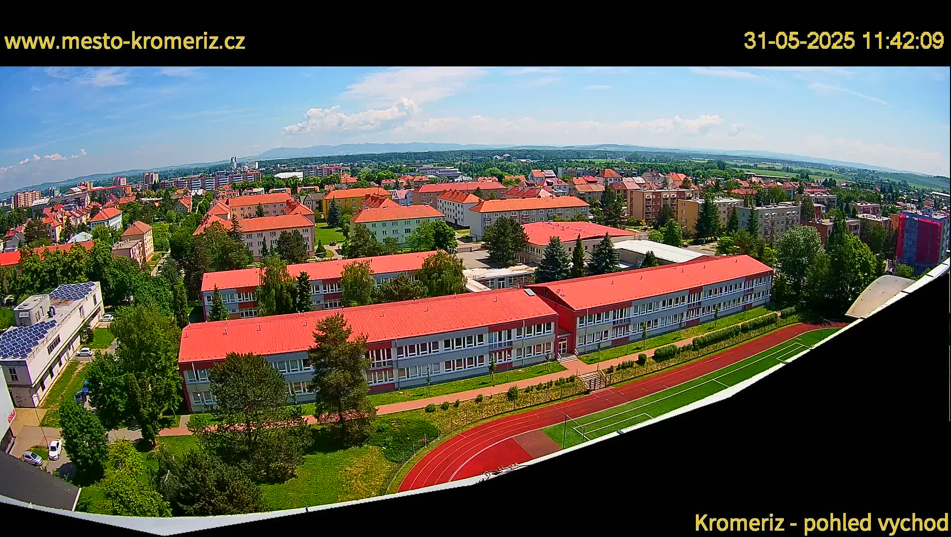 An aerial view shows a school with red roofs and a running track, surrounded by residential buildings and lush greenery under a partly cloudy sky.