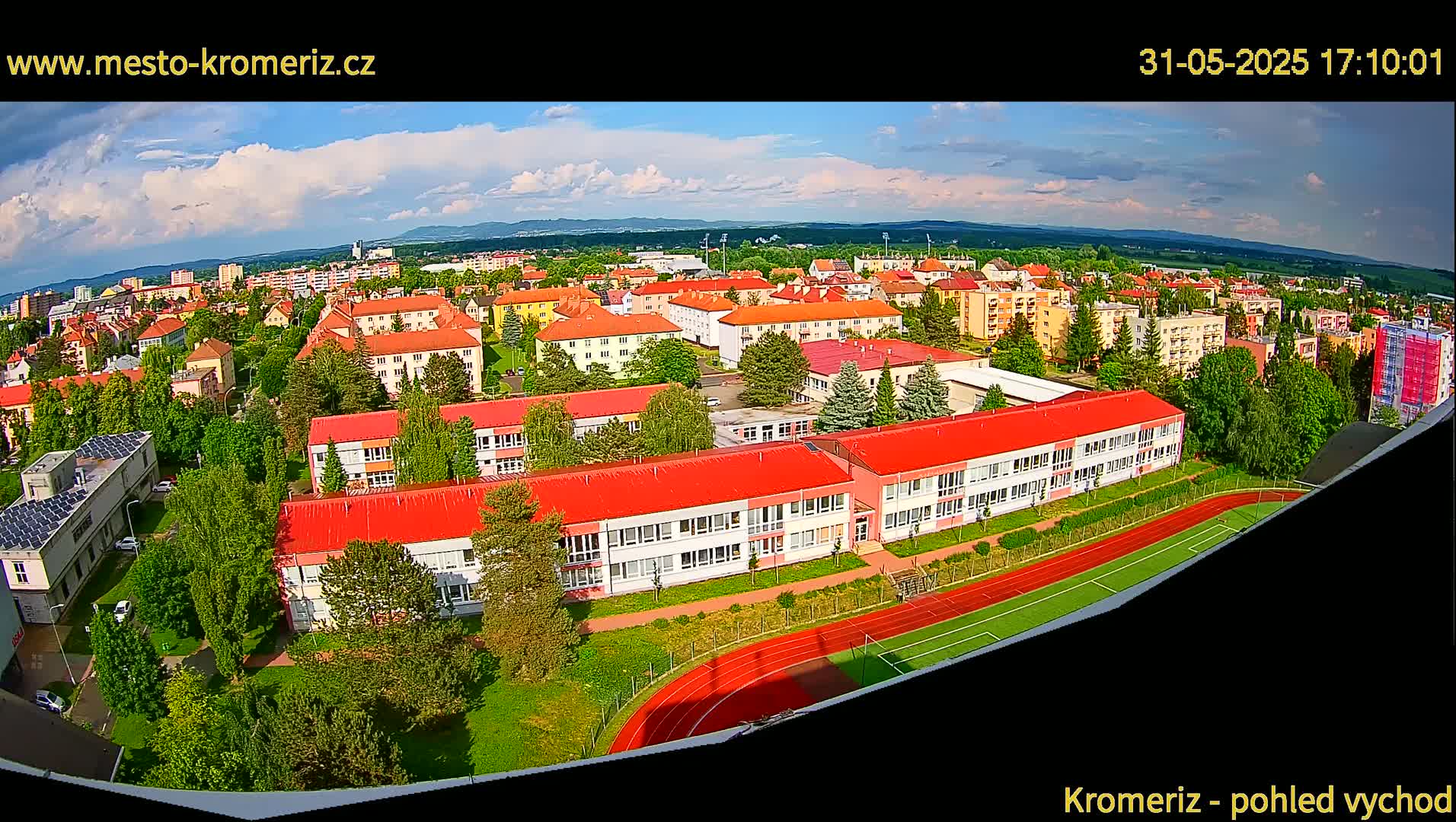 A partly cloudy daytime aerial view shows a large school building with red roofs and a running track, surrounded by other buildings and green trees in a residential area.