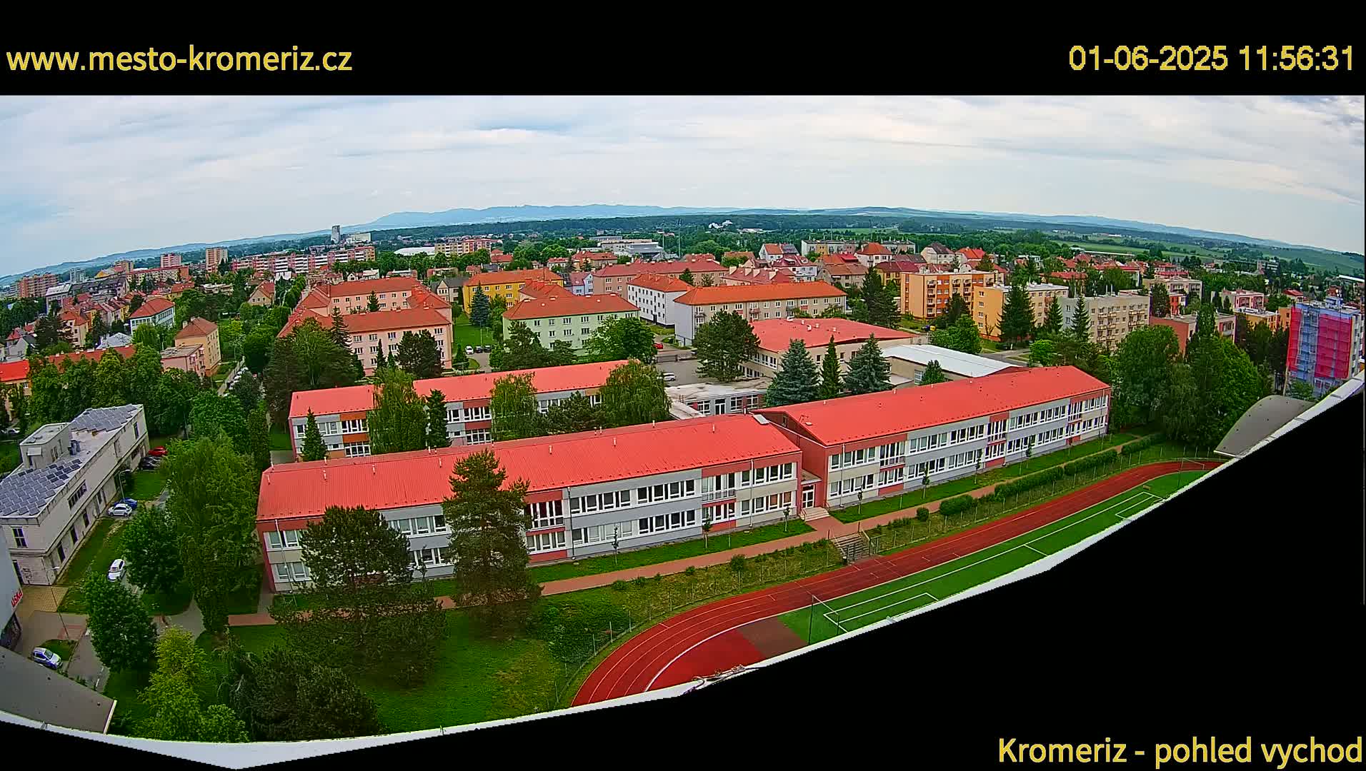 A high-angle view of a town under partly cloudy skies shows several buildings, including a large school with a red roof and a running track, surrounded by green spaces.