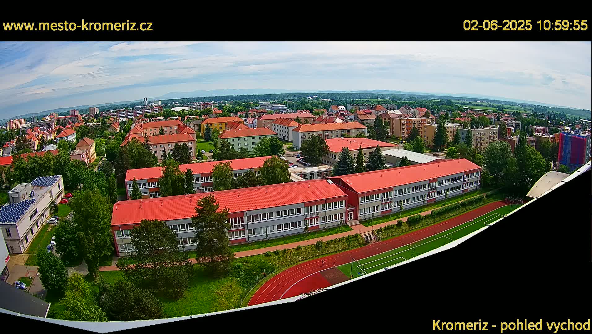 An aerial view shows a town under partly cloudy skies, with a large school building and athletic field prominently featured.