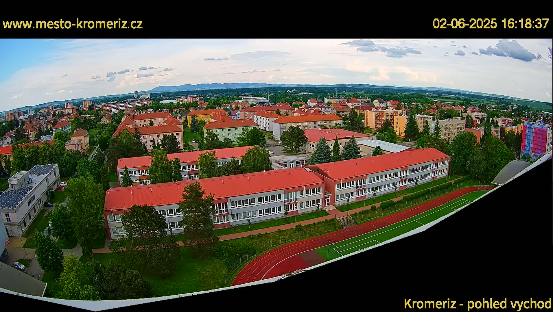 An aerial view of a town under partly cloudy skies shows numerous buildings, including a large school with a running track and soccer field, surrounded by green trees and landscaping.
