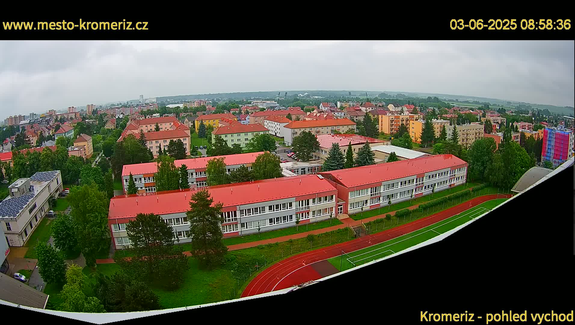 An aerial view shows a town under an overcast sky with numerous buildings, including a large school with a red roof and a running track, surrounded by greenery.