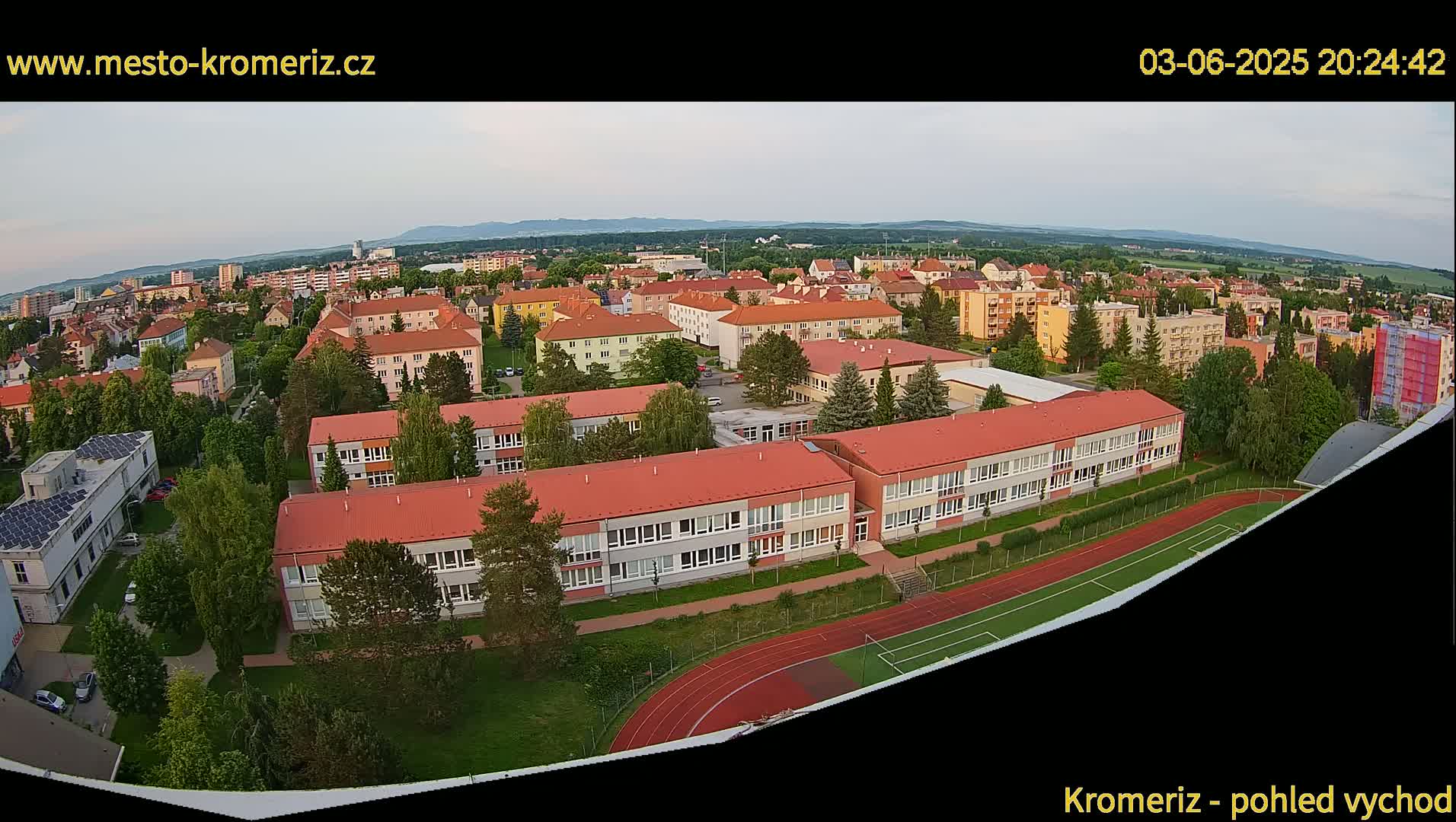 An aerial view shows a residential area with several school buildings and a running track under a partly cloudy evening sky.