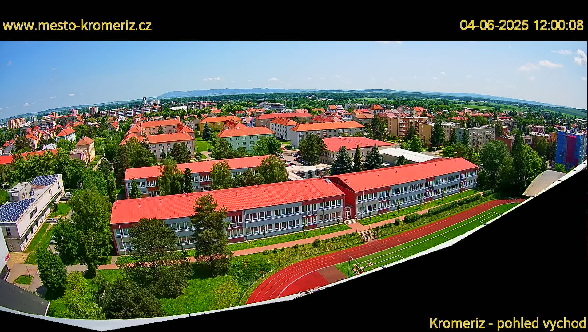 An aerial view shows a large school complex with red roofs, surrounded by green lawns and trees, next to a running track and a playing field, on a clear, sunny day.