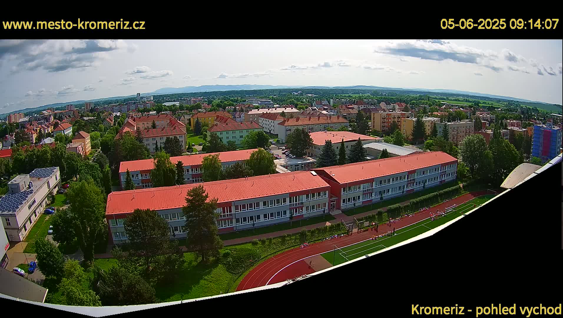 An aerial view shows a town on a partly cloudy day, with a large school building and a running track visible in the foreground.