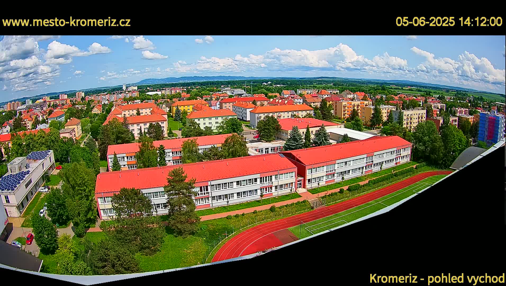 A partly cloudy day reveals a panoramic view of a town with numerous buildings, including a large school with a red roof and a running track, surrounded by green spaces.
