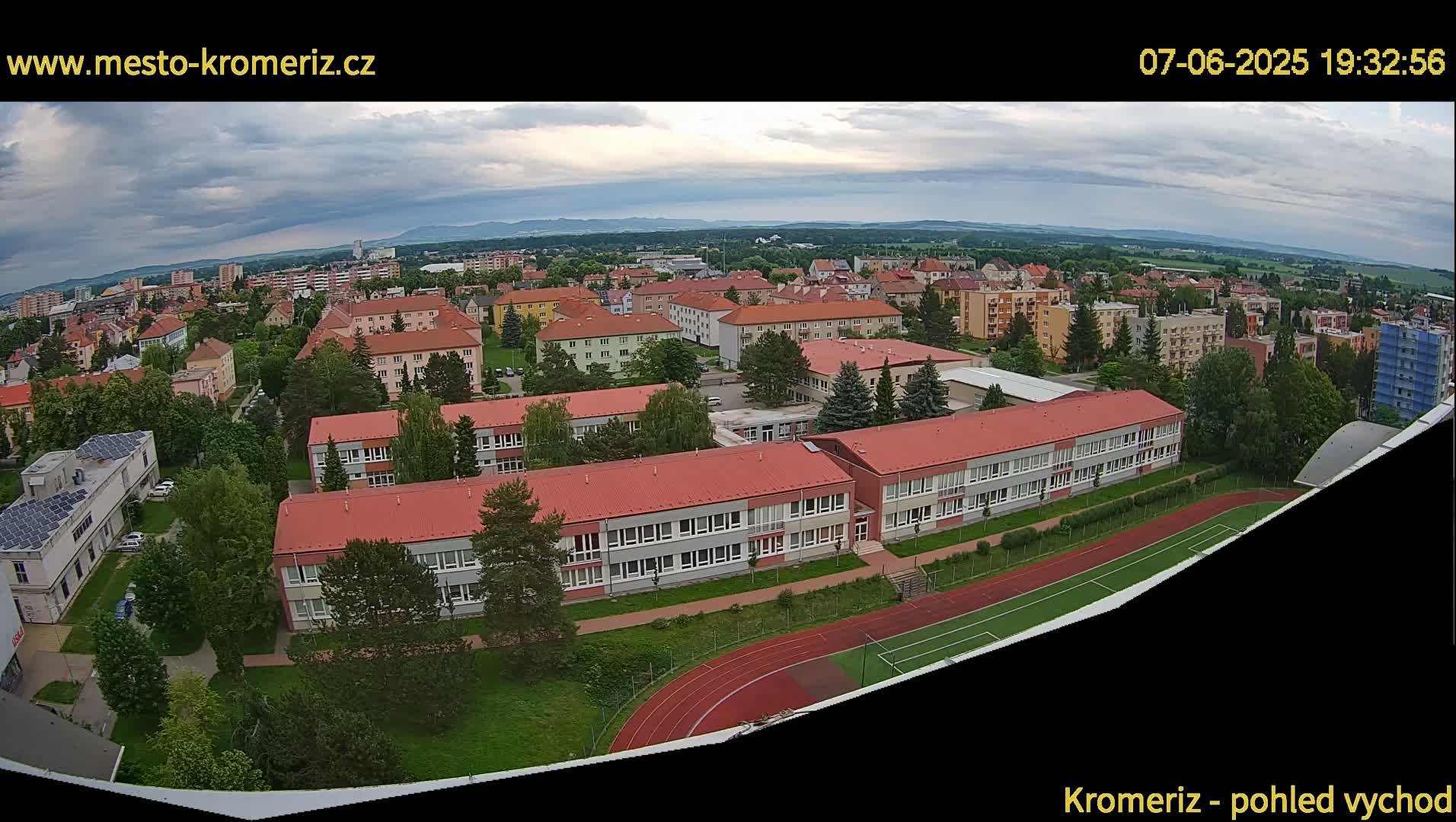 An aerial view of a town under partly cloudy skies shows a large school building with a red roof and adjacent athletic fields.