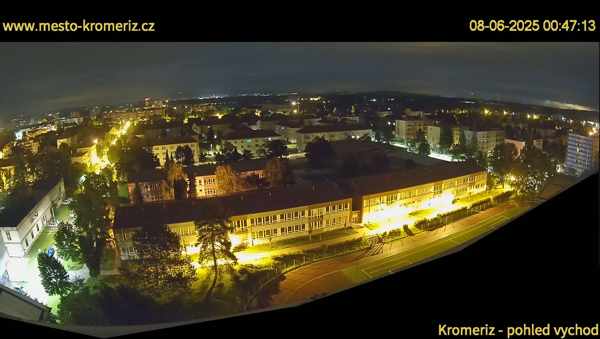 A nighttime aerial view of a town shows illuminated buildings, streets, and a sports field.