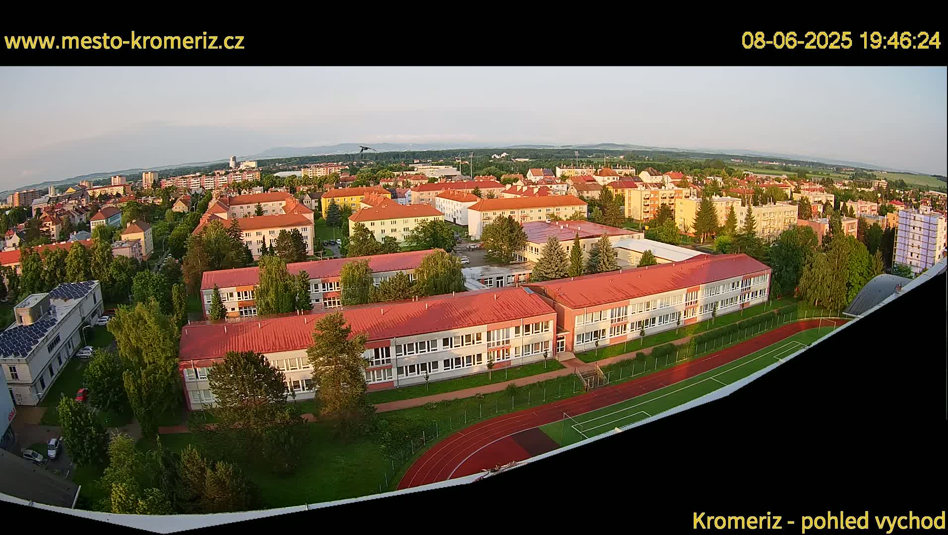 An aerial view shows a large school building with red roofs and a running track, surrounded by other buildings and trees under a clear evening sky.