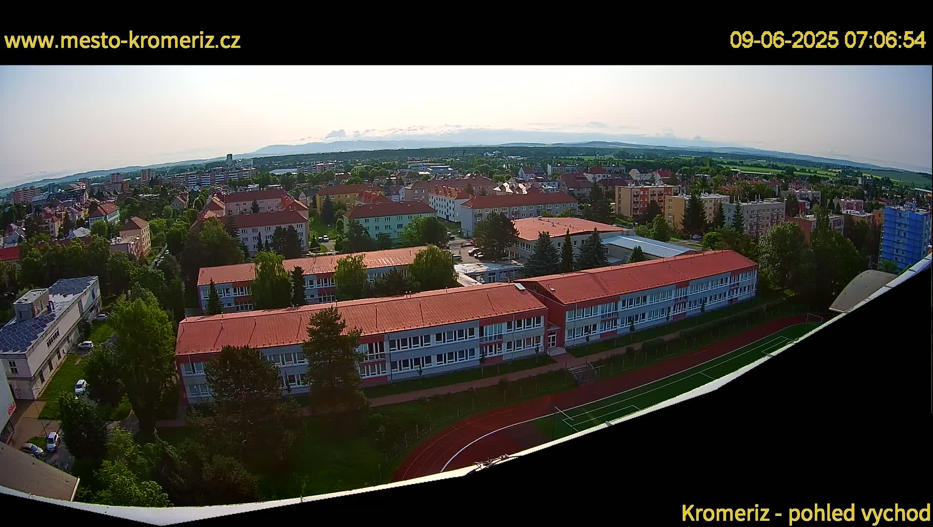 An aerial view shows a town on a partly cloudy day with a large school building and surrounding residential areas, a running track, and distant mountains.