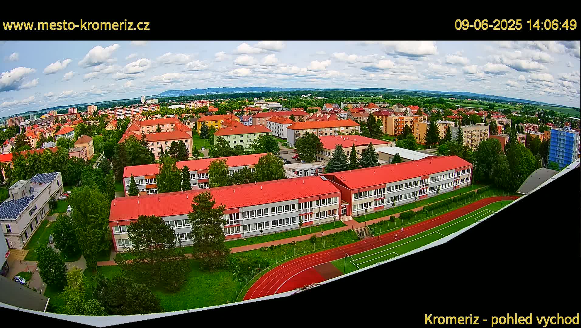 An aerial view shows a school complex with red roofs and a running track on a partly cloudy day.
