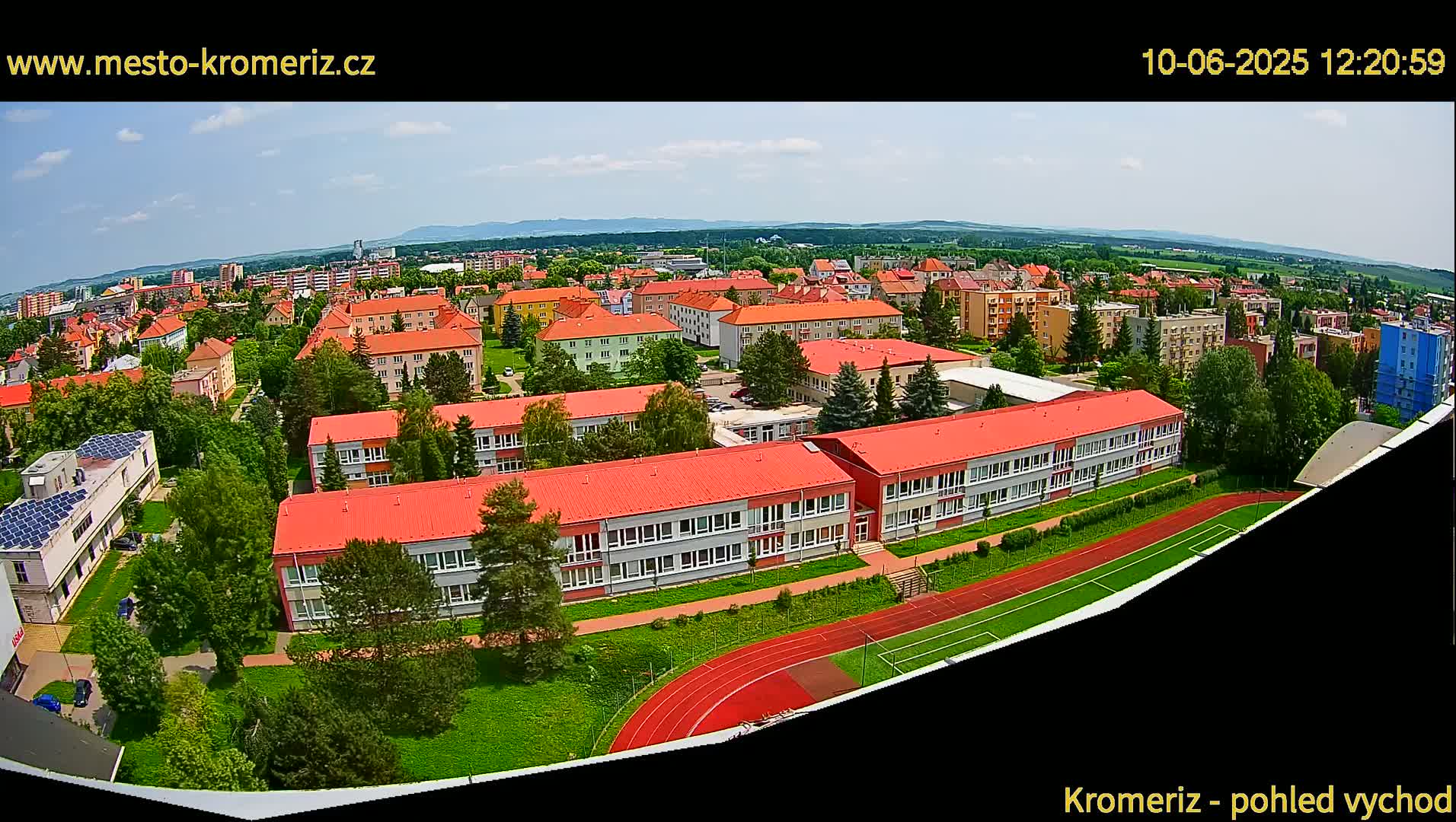 An aerial view shows a large school complex with red roofs surrounded by green landscaping and a running track, on a partly cloudy day.