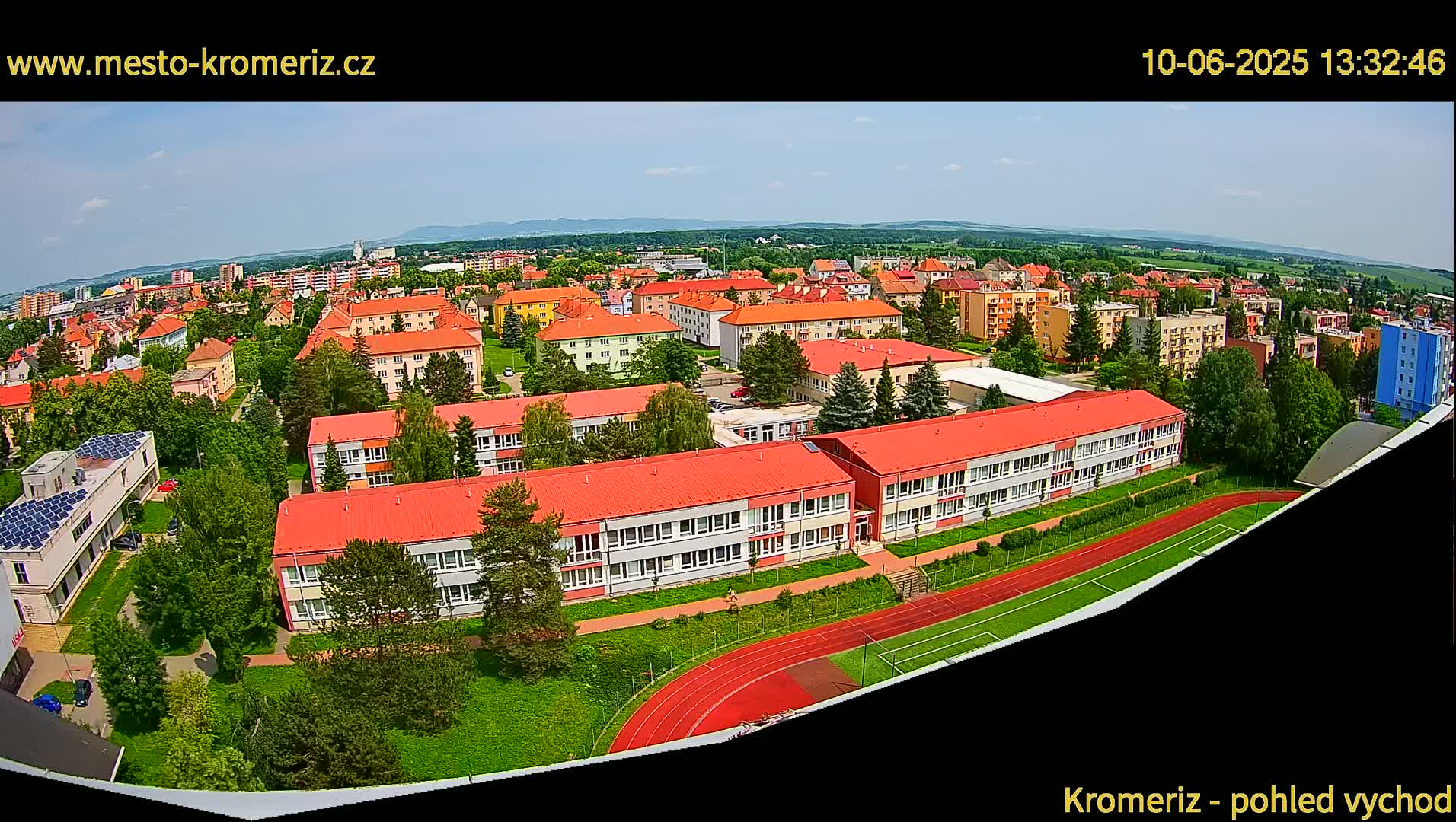 A wide, high-angle view shows a large school complex with red roofs, surrounded by green landscaping and a running track, situated within a residential area on a clear, sunny day.