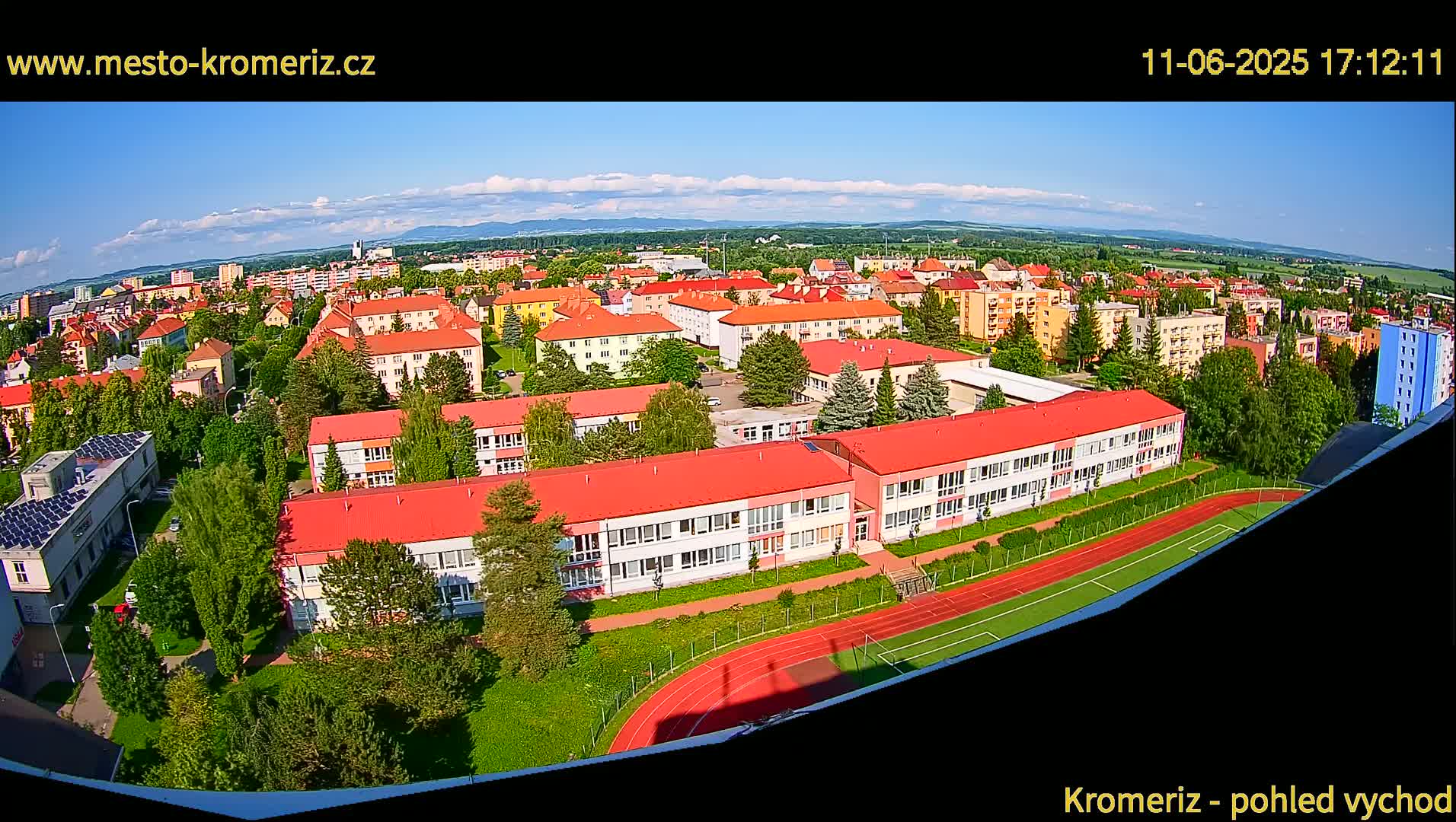 An aerial view of a town on a partly sunny day shows a large school building with red roofs and a running track next to it, surrounded by other buildings and green spaces.