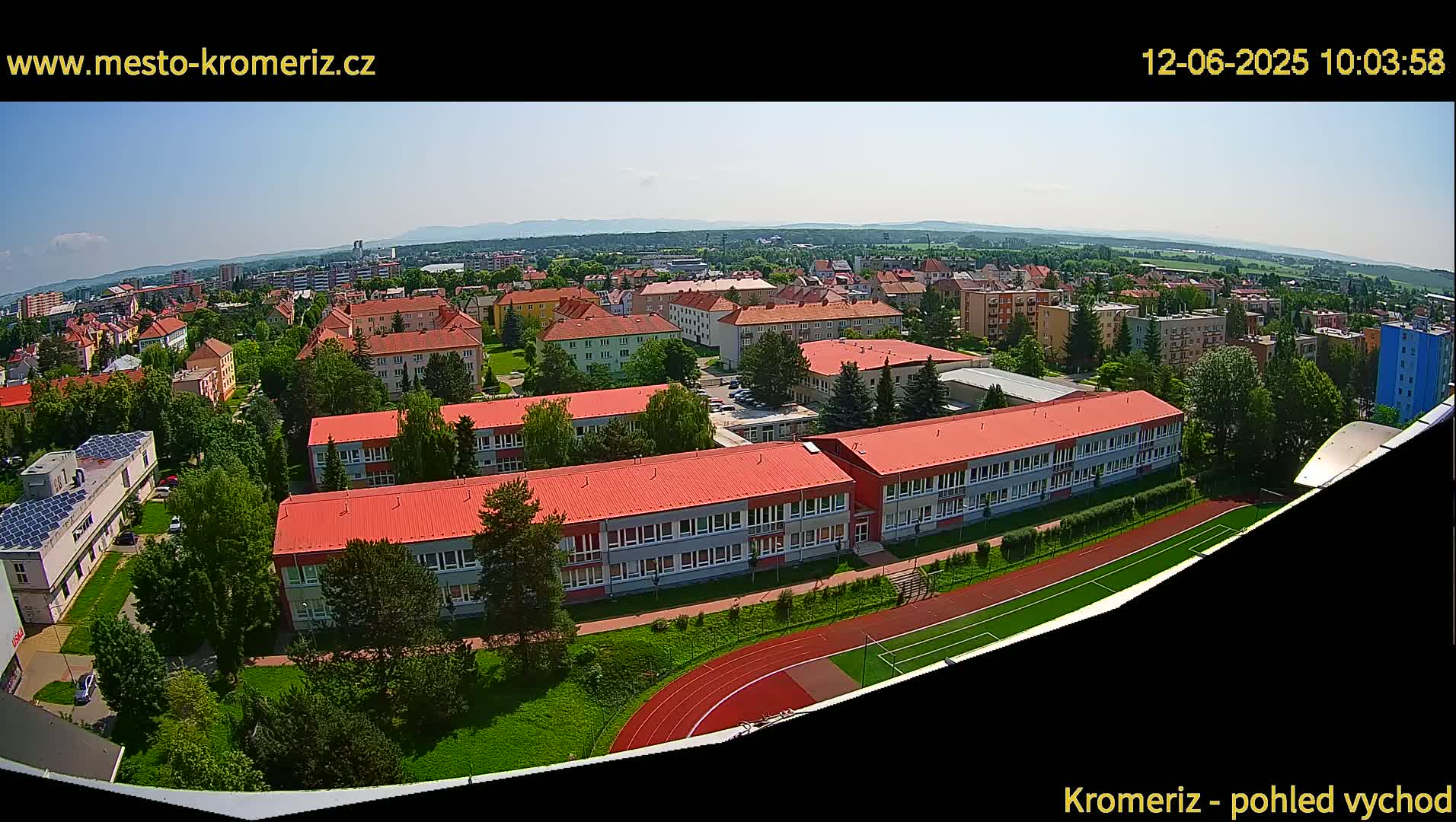 An aerial view of a town on a sunny day, showing a large school building with red roofs and a running track, surrounded by other buildings and green spaces.