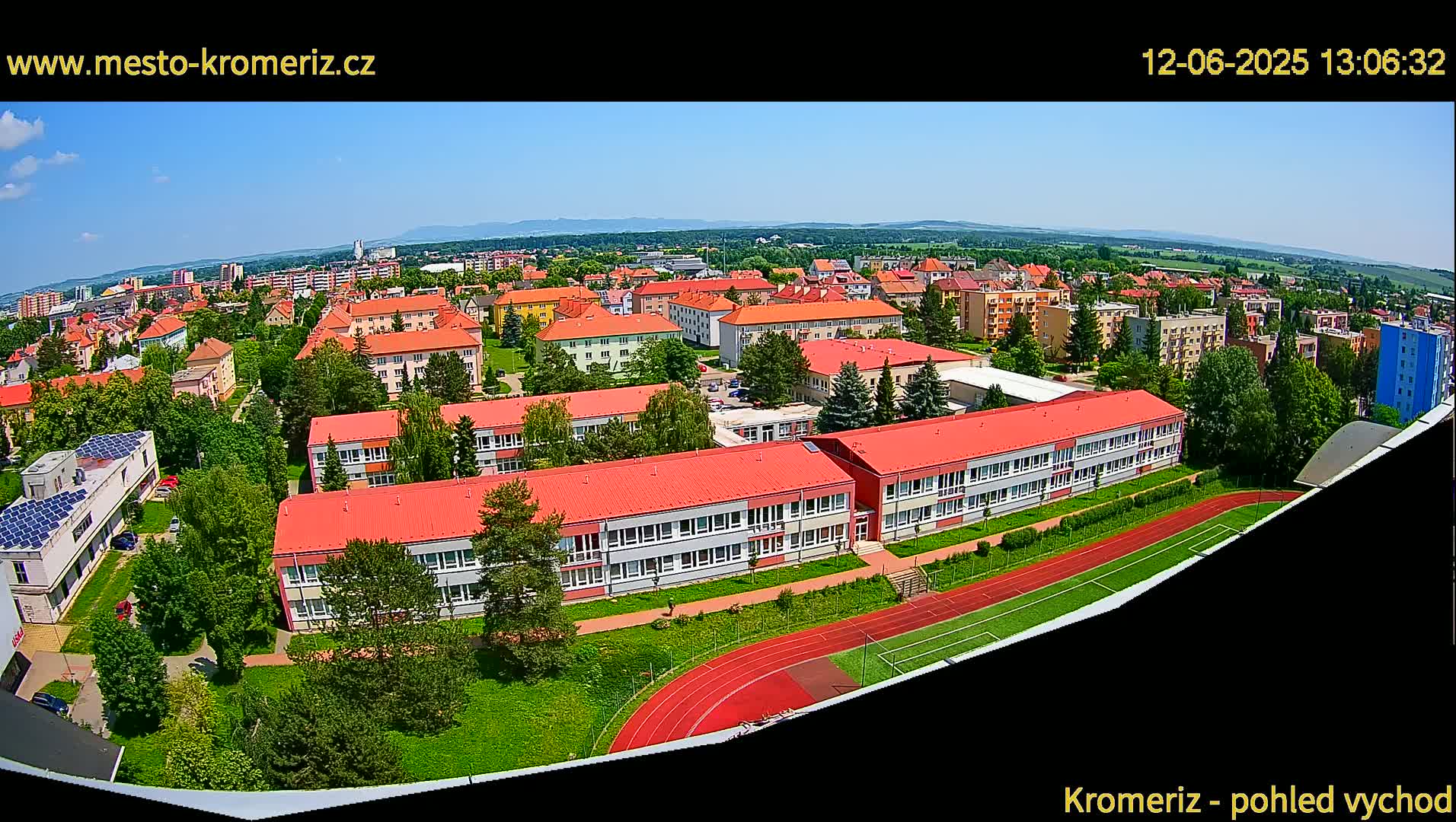 An aerial view shows a town under a clear blue sky, featuring several buildings with red roofs, a running track, and surrounding green areas.