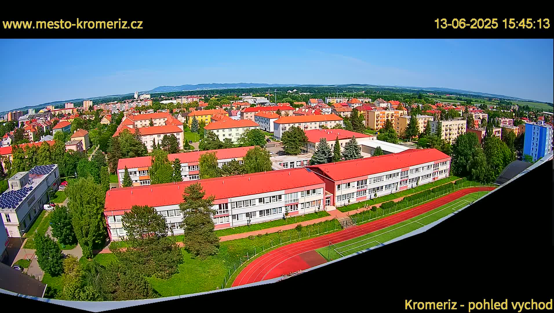 An aerial view of a town on a sunny day, showing a large school building with a red roof and a running track next to it.