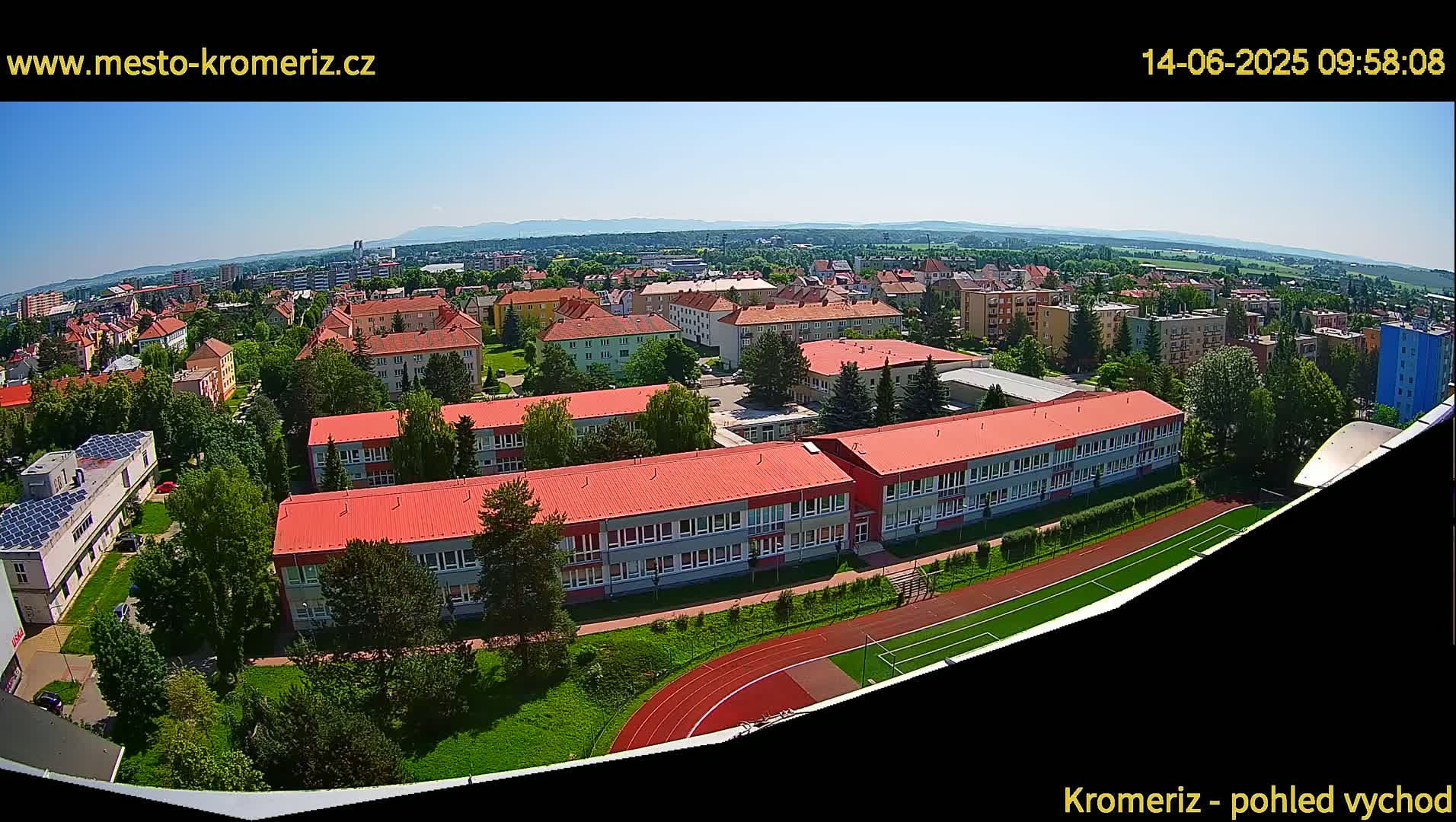 An aerial view on a sunny day shows a large school building with a red roof and a running track, surrounded by other buildings and green spaces.