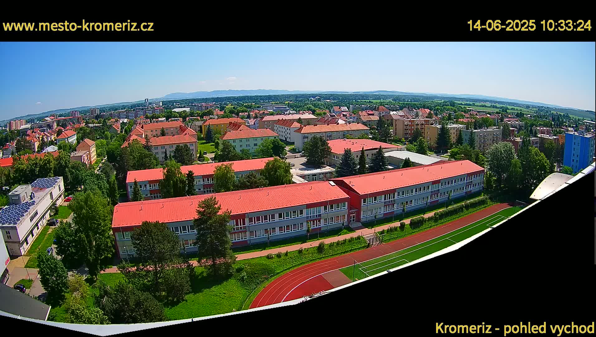 A wide aerial view shows a school complex with red roofs and a running track on a sunny day, surrounded by residential buildings and greenery, with mountains visible in the distance.