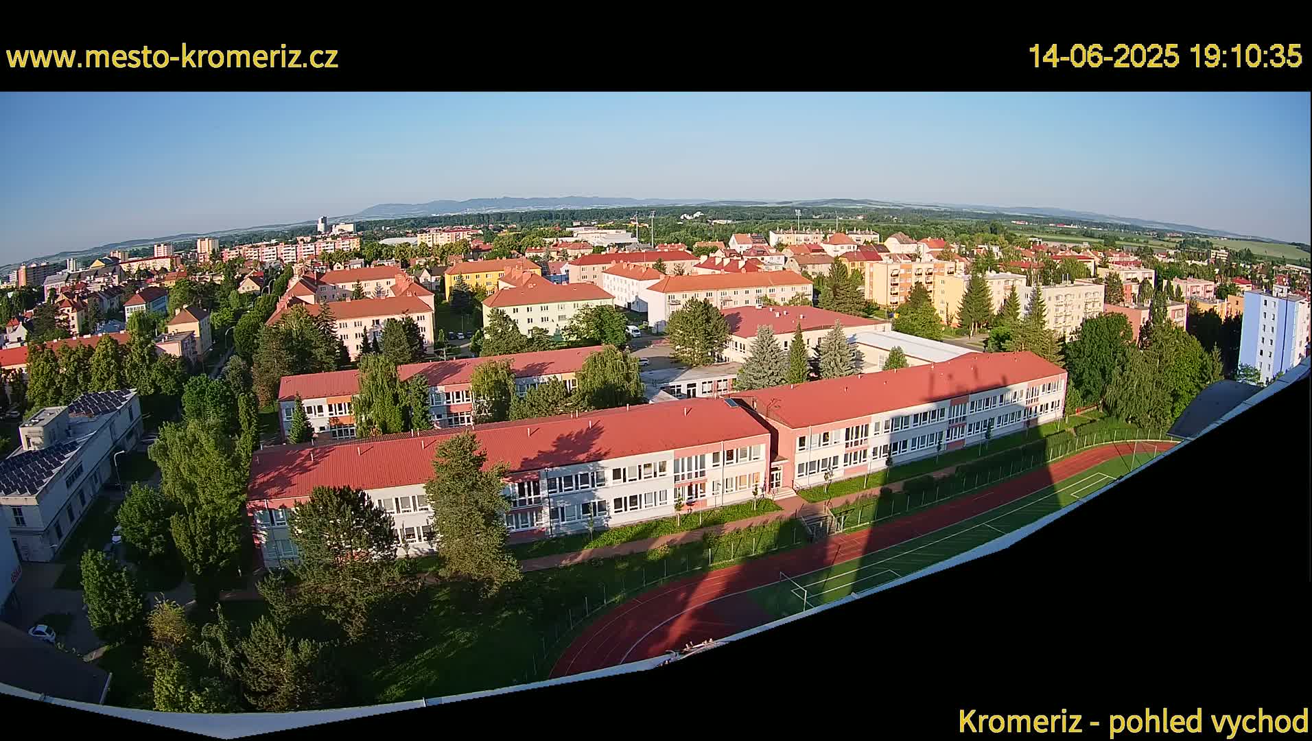 A wide aerial view shows a school complex with red roofs and a running track on a sunny day, surrounded by residential buildings and greenery, with mountains visible in the distance.
