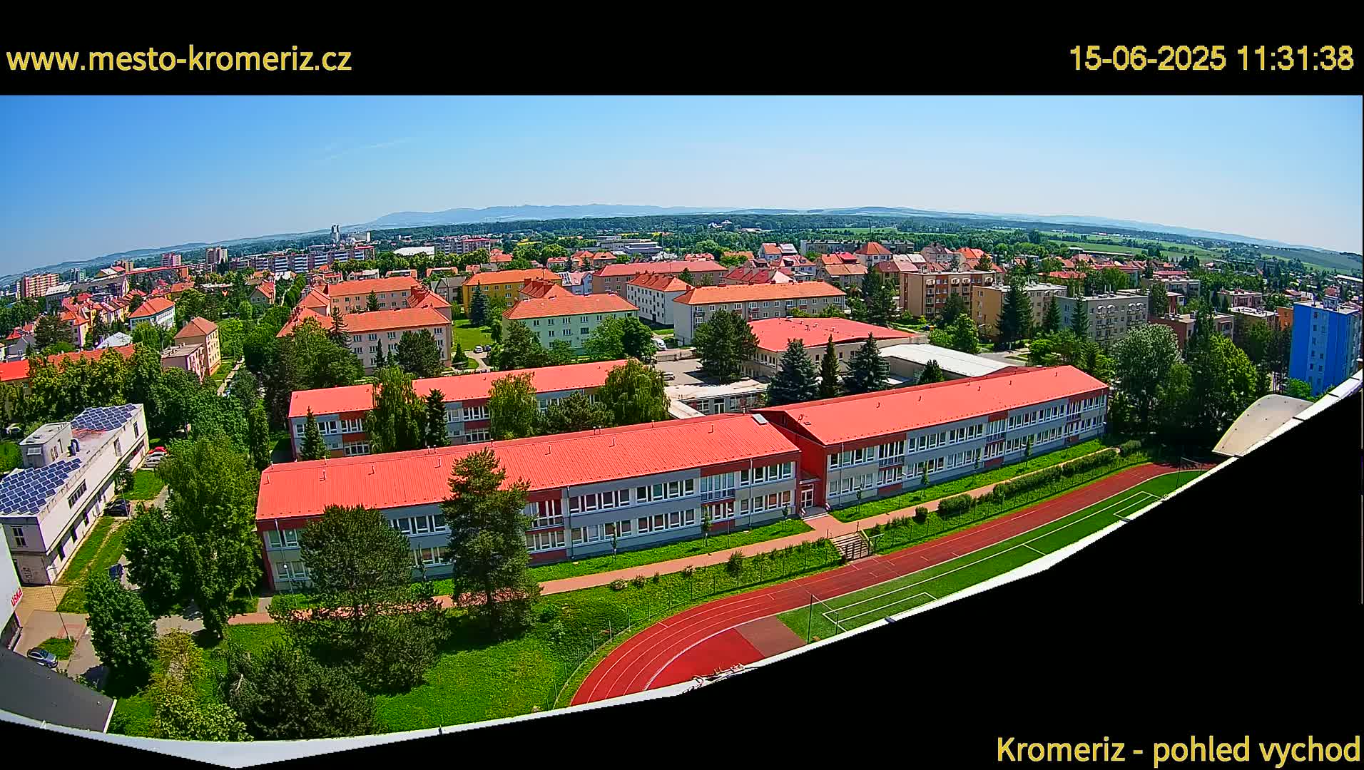 An aerial view on a sunny day shows a large school complex with red roofs and a running track, surrounded by other buildings and green spaces.