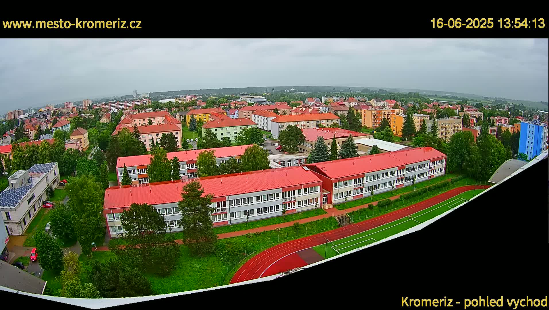 An aerial view of a town on an overcast day shows several buildings with red roofs, a running track, and green spaces.
