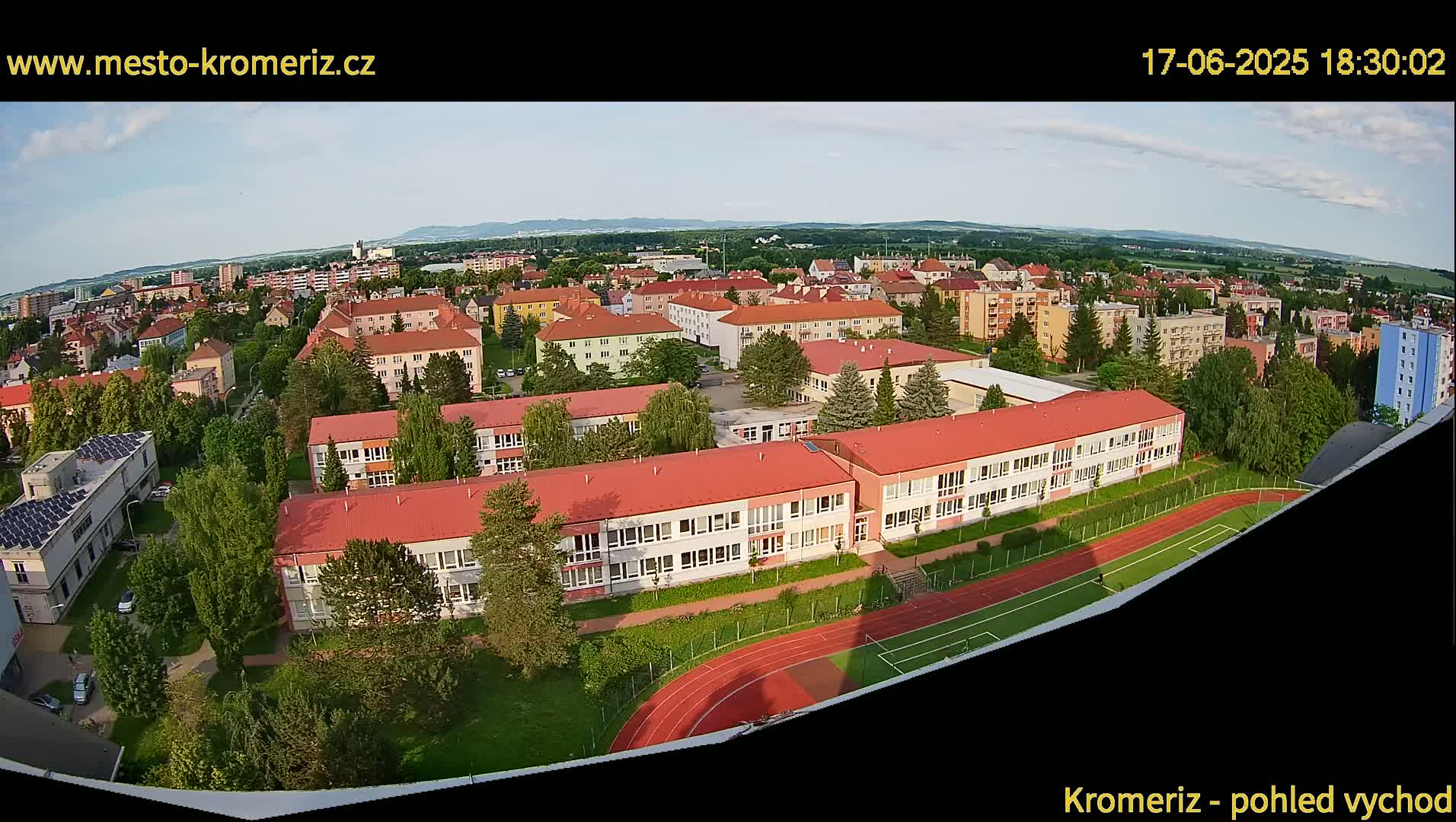 An aerial view shows a residential area with several school buildings, a running track, and green spaces under a mostly clear sky.