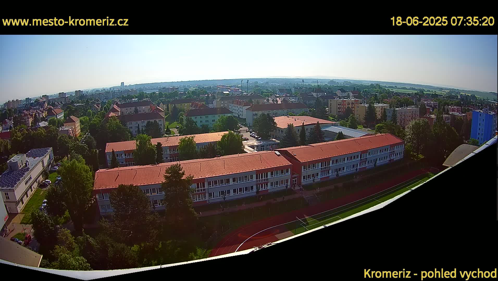 An aerial view of a town on a clear, sunny day shows a large school building with a red roof and a running track next to it, surrounded by other buildings and trees.