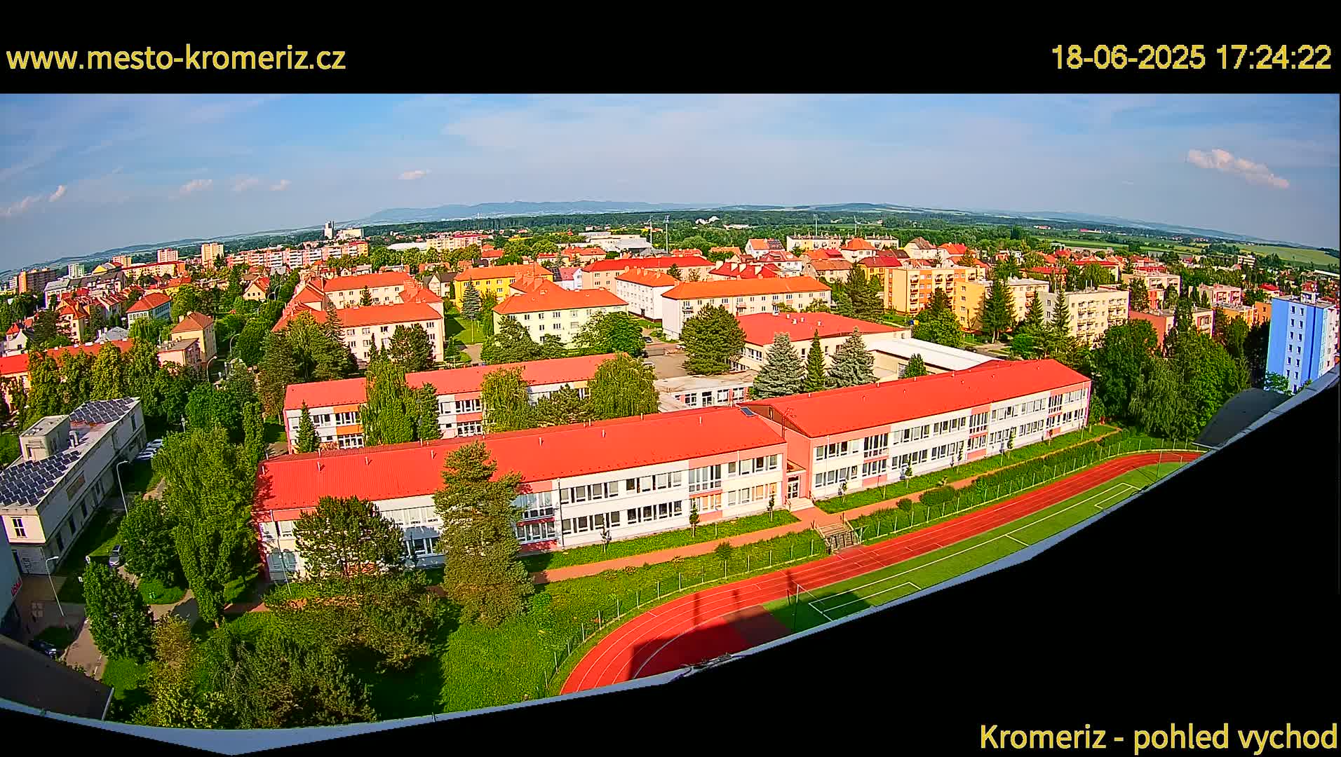 An aerial view shows a town on a partly sunny day, with a large school building and a running track prominently featured in the foreground.